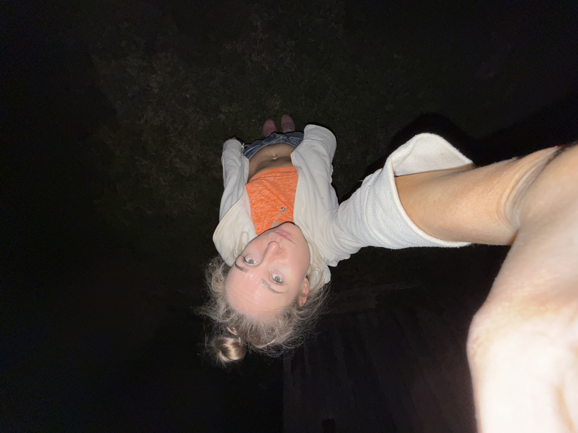 A woman stands outside at night in a dark area. She is fair-skinned with blonde hair in a bun. She wears an orange shirt, jeans, and a white hoodie. She is looking upwards at the camera, which is held at arm's length. The location is unknown.