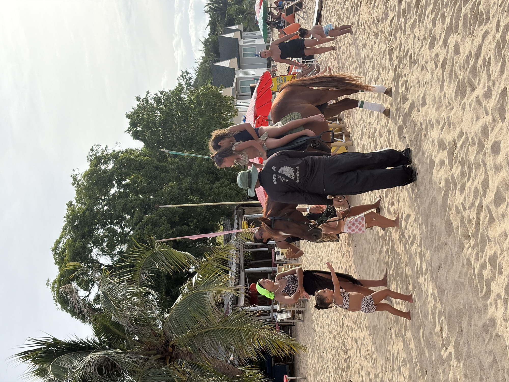 The image depicts a sunny day at a beach in Ko Samui District, Thailand. The scene features a brown horse on a sandy beach with several people around it. Two young girls are positioned on the horse's back, one appearing to be riding and the other close behind her. A man in dark clothing is standing next to the horse, holding its reins and appearing to guide it. Further back, other individuals are visible in the background, some seated on beach chairs under umbrellas, and others standing. Tropical trees and vegetation are present to the left of the frame, and in the far background, several white buildings with dark roofs suggest a resort or hotel area. The sand is well-lit, indicating daytime. One of the visible signs on the man's shirt has text in what appears to be Thai. The overall atmosphere suggests a leisurely day at a tourist destination, with horse riding being an activity offered.