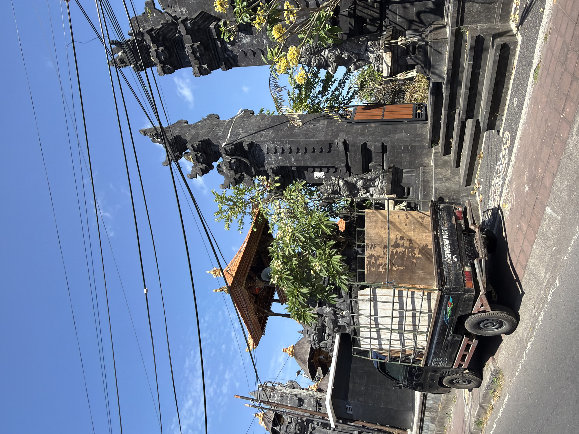 The image depicts a street scene in Kuta Selatan, Indonesia, featuring a black pickup truck parked on the side of a road. The truck has a cargo bed filled with materials, including wooden planks and what appears to be metal scaffolding. In the background, a large, ornate black stone gateway with intricate carvings and statues stands prominently. This gateway, typical of Balinese architecture, is adorned with traditional roofing and is flanked by lush greenery. Overhead, a complex network of electrical wires crisscrosses the bright blue sky, indicating an urban or semi-urban environment. The ground is paved with textured bricks and concrete, with some fallen leaves scattered about. The overall impression is of a bustling, culturally rich locale under a clear, sunny sky.