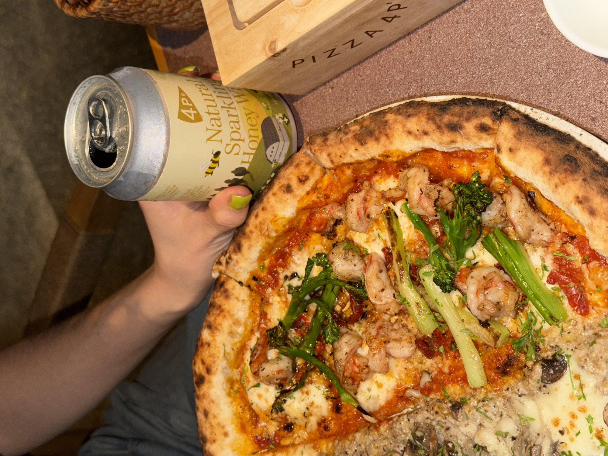 A partially consumed pizza sits on a white plate, positioned adjacent to a light brown wooden box inscribed with "PIZZA 4P". A light green aluminum can of "4P's Natural Sparkling Honey Water" is held in a hand with light green nail polish. The pizza features a charred crust, with visible toppings including shrimp and green vegetables. The image is taken indoors, possibly in a restaurant or dining area in Đà Nẵng, Vietnam.