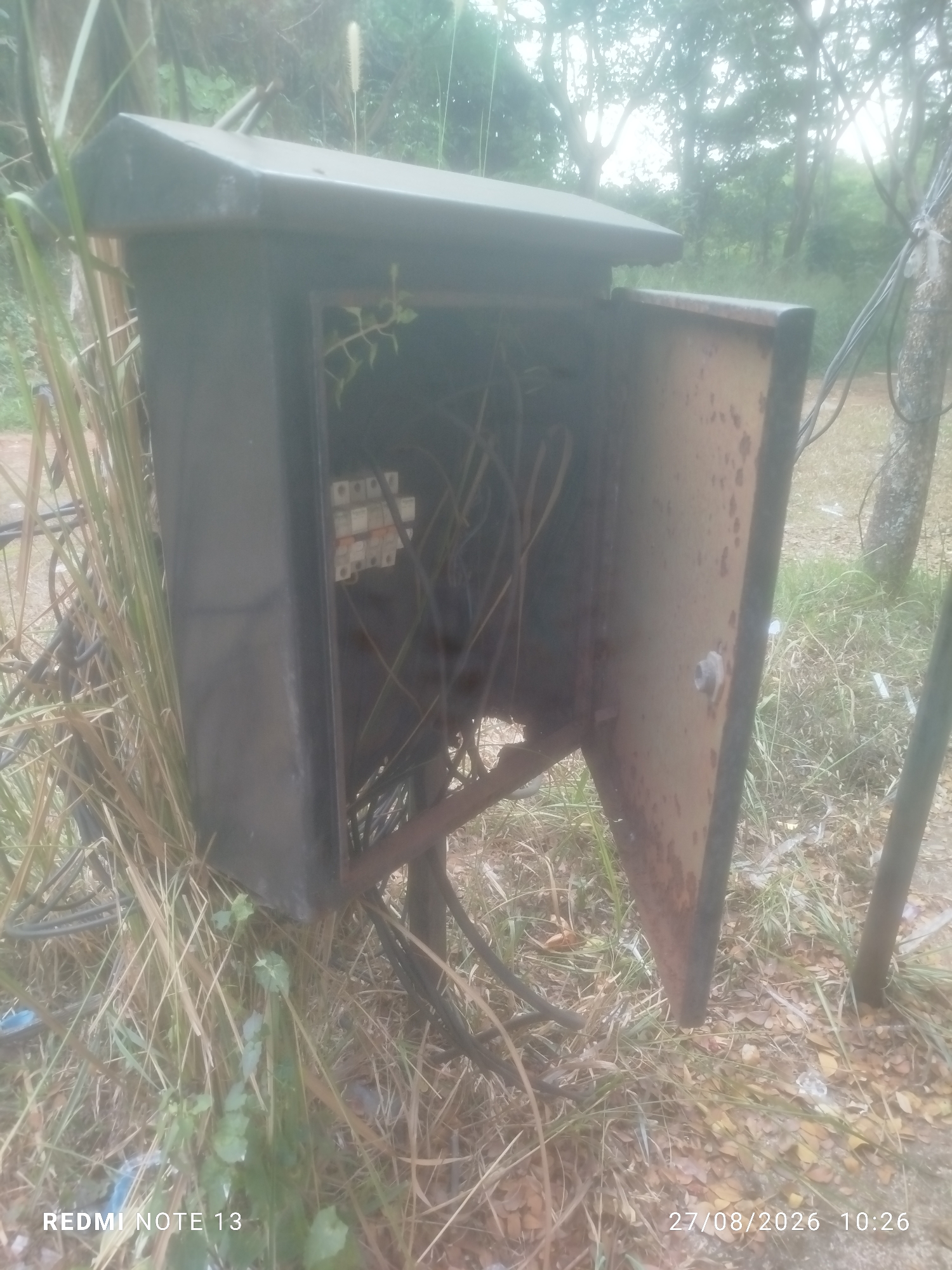 An outdoor, slightly overcast scene shows a rusted, dark metal electrical junction box with its right door ajar, revealing a tangle of wires and some small circuit breakers within. The box is mounted on a metal stand and is surrounded by tall, dry grass and fallen leaves, suggesting an unmaintained or rural area. In the background, trees and foliage create a dense, green environment. The image is watermarked with "REDMI NOTE 13" and "27/08/2026 10:26" in the lower corners.