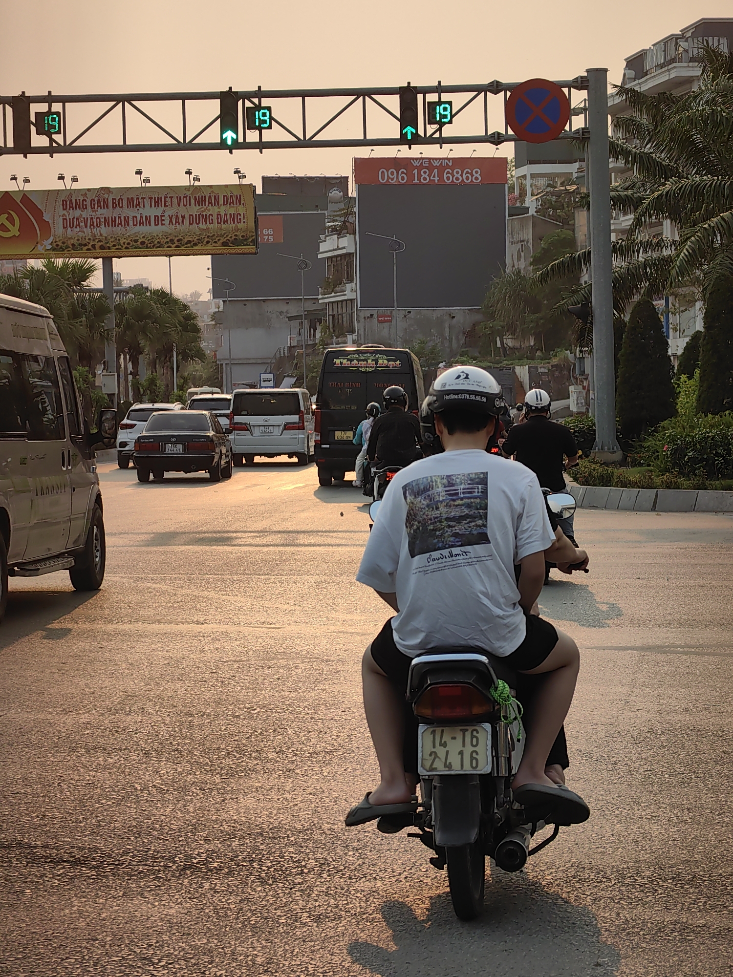 The image captures a street scene in Hong Gai Ward, Vietnam, during daylight. A motorcyclist, wearing a white t-shirt with a print and helmet, is seen from behind, riding on the road. On the back of the motorcycle, the license plate reads "14-T6 2416". A passenger is also on the motorcycle. Ahead, a line of vehicles, including cars and vans, are moving in the same direction. Other motorcyclists are also visible, further ahead. Above the road, there is a traffic light indicating 19 seconds. A sign with the text "WE WIN 096 184 6868" and another banner with Vietnamese text are visible. Palm trees and buildings line the road.
