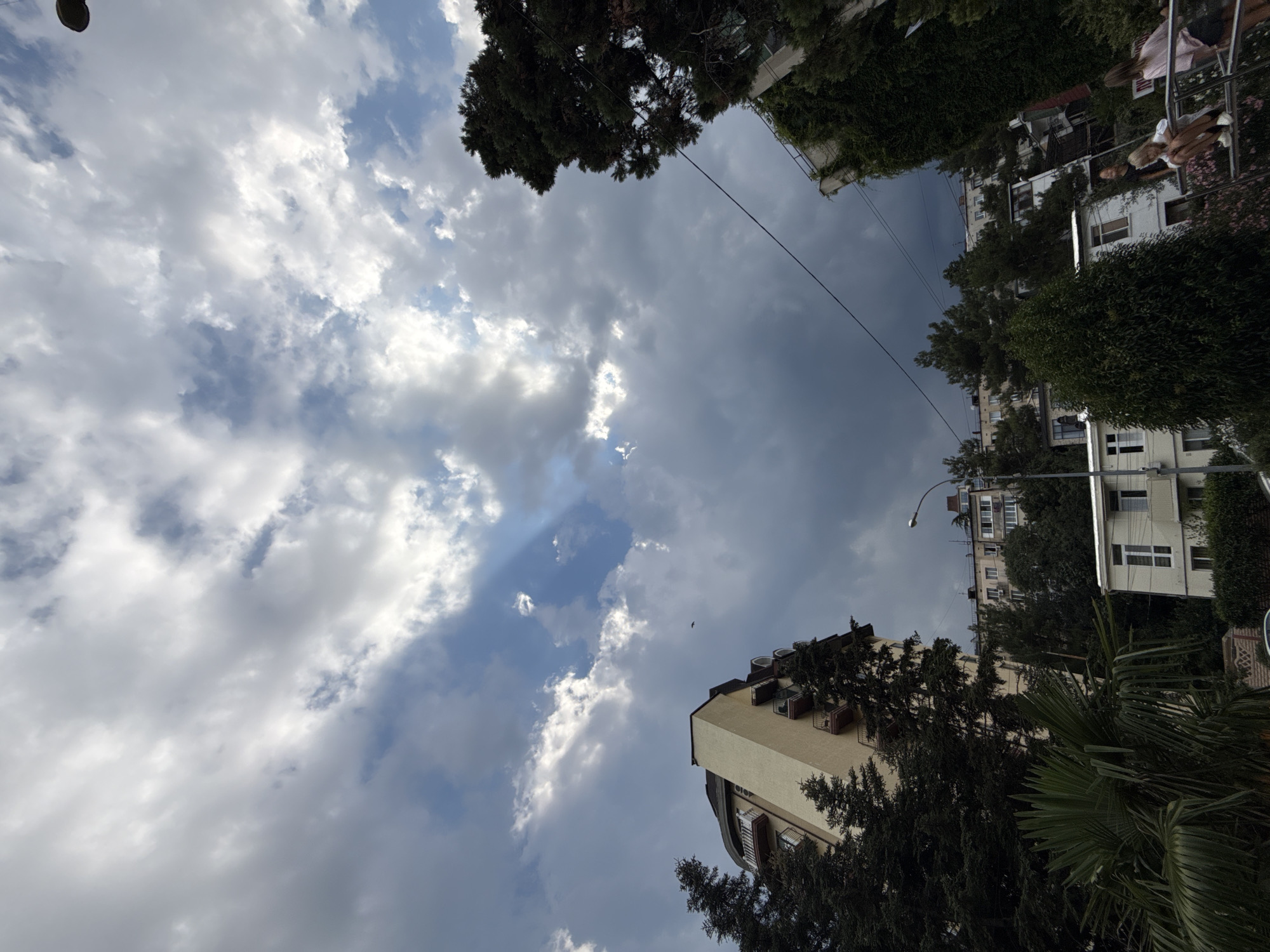 In Partenit, Ukraine, the sky above a residential area is filled with dramatic clouds, with rays of sunlight breaking through in the upper left. A distinct, curved building with a pale yellow facade stands out on the left, surrounded by tall, dark green evergreen trees. In the background, several other apartment buildings are visible, nestled amongst more trees. A lamppost is situated above a white, multi-story building in the mid-ground. On the right side of the image, a railing is visible with two young females, one sitting and one standing, interacting on what appears to be a playground or outdoor seating area. Lush greenery and flowering plants contribute to the urban environment.