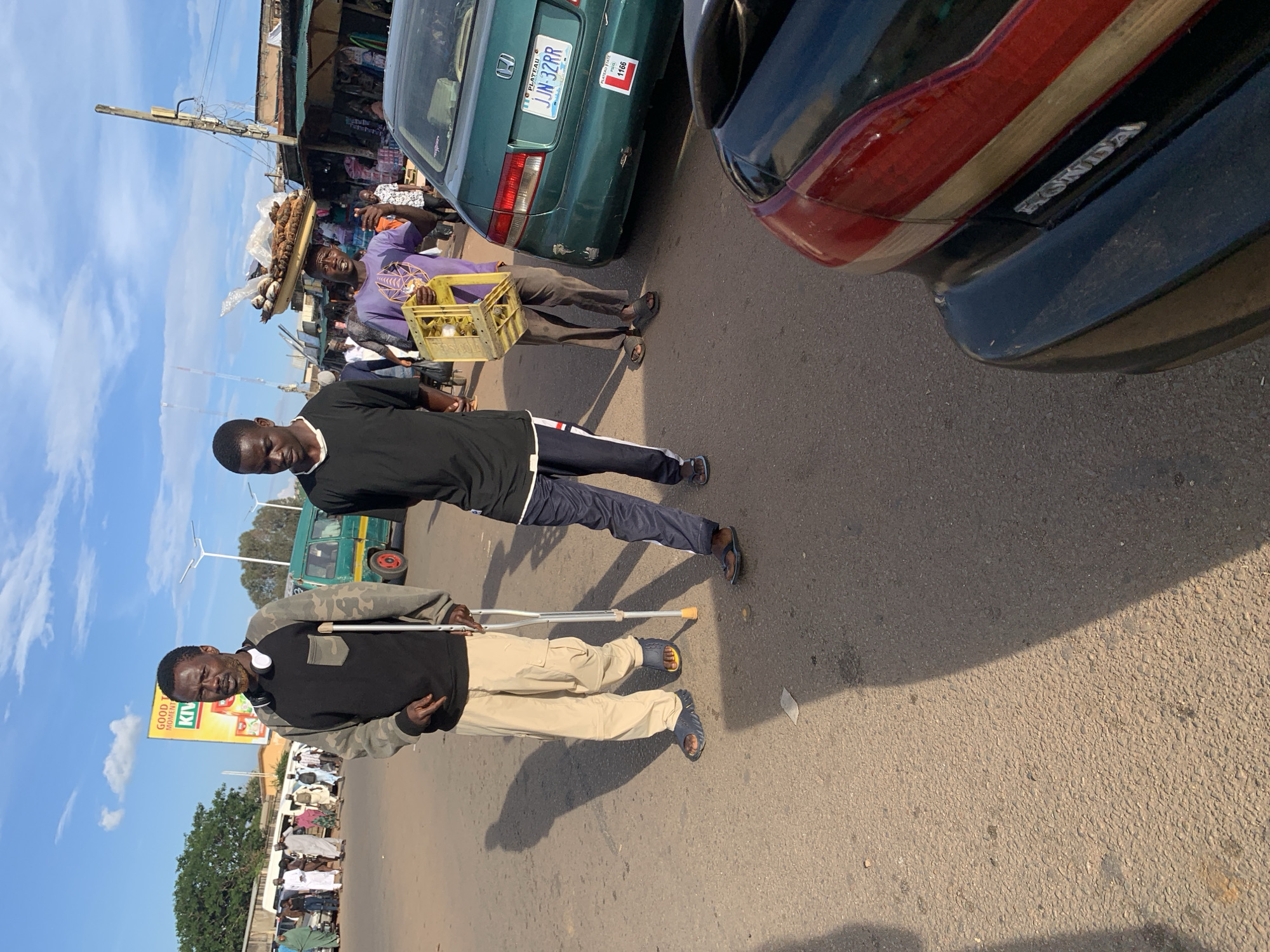 The image depicts a street scene in Jos, Nigeria, on a sunny day. In the foreground, a person with crutches walks with another individual. The person with crutches wears a camouflage-patterned sweatshirt and khaki pants. To their right, another person in a black t-shirt and dark pants walks by. In the middle ground, a vendor balances a tray of fish on their head, holding a yellow crate. Behind them, a green sedan with a Nigerian license plate is parked. Further in the background, several people are seen near market stalls and a yellow bus. The sky is blue with scattered clouds, and the overall environment suggests a busy urban area.