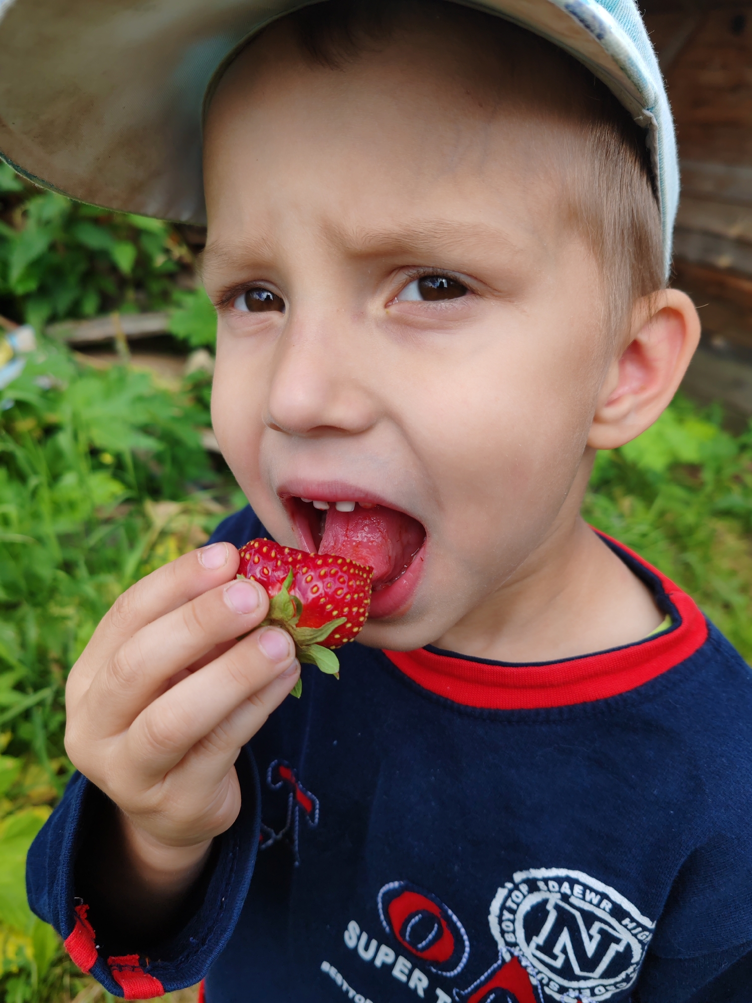 A young boy, appearing to be of Caucasian ethnicity and possibly between 4-6 years old, is shown in a close-up portrait as he eats a strawberry. He is wearing a navy blue long-sleeved shirt with a red trim around the collar and a light-colored baseball cap. His mouth is open as he bites into the strawberry, which he holds in his right hand. The background consists of green foliage and what appears to be a wooden structure, suggesting an outdoor setting, possibly a garden or rural area. The overall lighting suggests daylight. There is no information to determine the specific location in the city of Чухлэм, Russia.