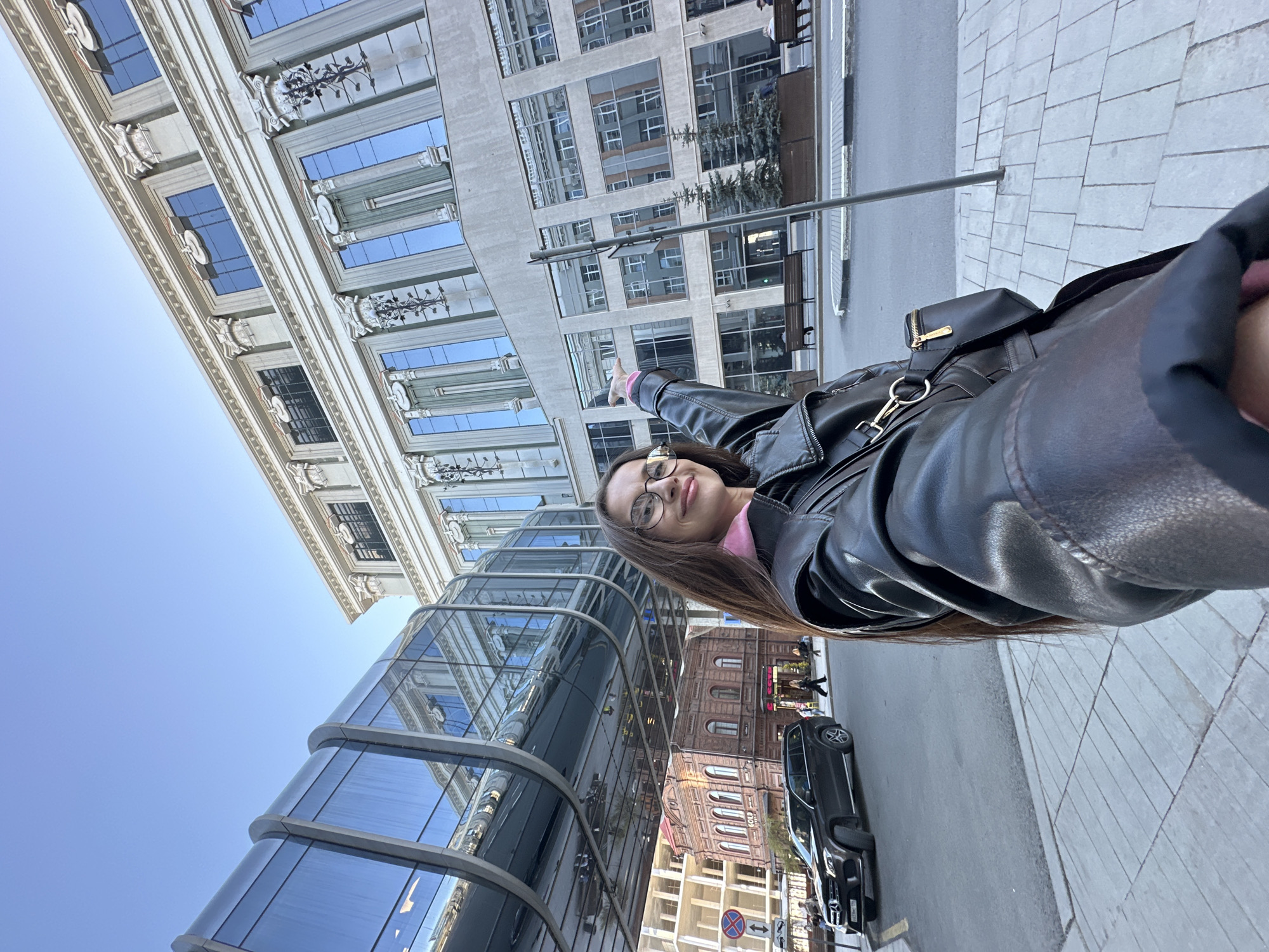 A person is standing in front of several buildings in Yekaterinburg, Russia. The person is wearing a black leather jacket and glasses, and pointing upwards with their left hand. A multi-story glass building extends to the left of the image, while older architectural styles can be seen in the background. A black car is parked on the street. The pavement is gray and made of rectangular tiles. There are other people visible in the background.