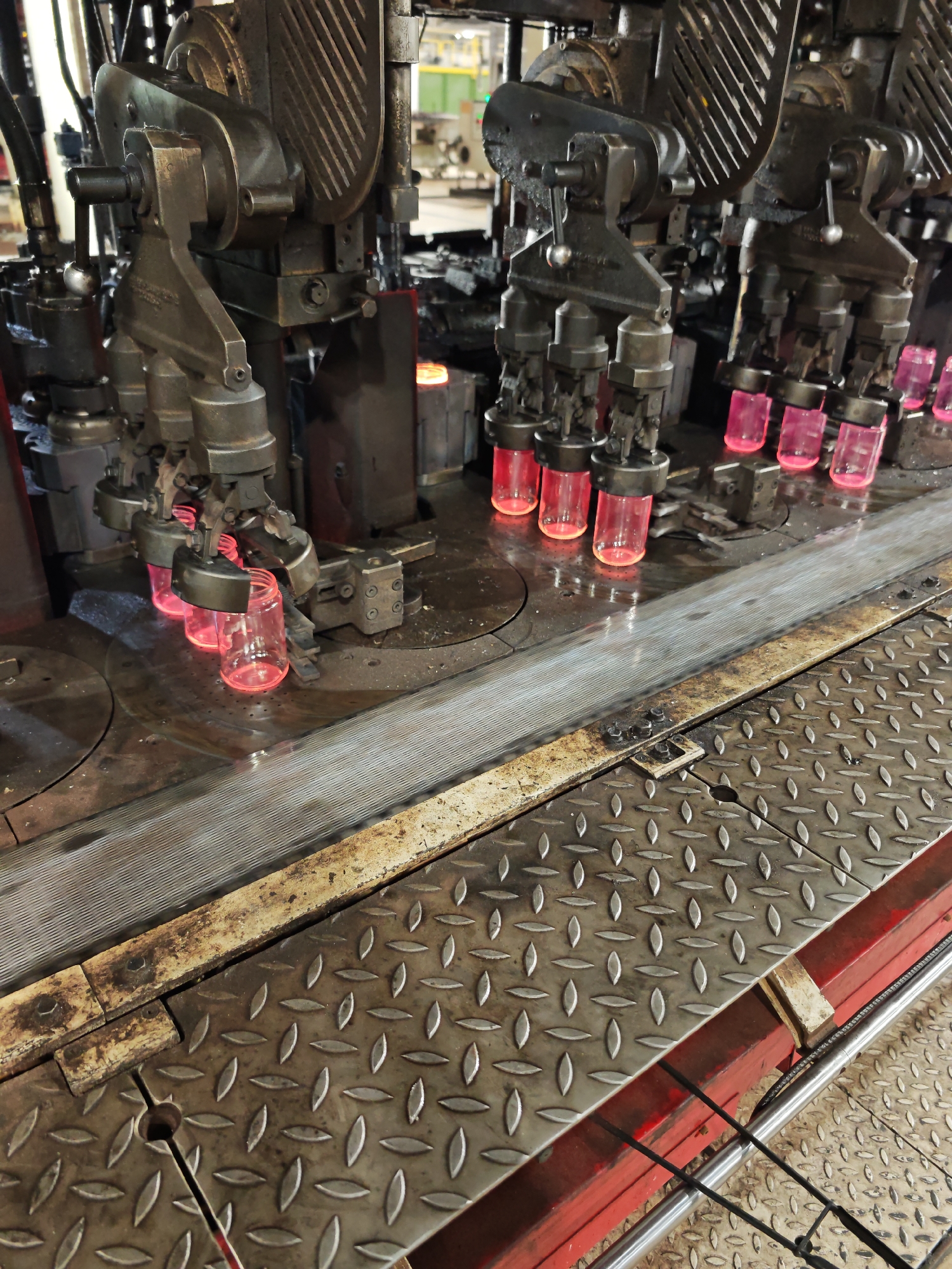 The image depicts a glass manufacturing process, likely in a factory setting. Molten glass is being shaped into jars or vials by an automated machine with multiple arms and forming tools. The newly formed glass containers are glowing red-hot. The machinery appears industrial and well-used, with signs of wear and grease. The floor is made of metal grating, suggesting a durable and practical environment. The specific location, Pleven, Bulgaria, is based on my knowledge of known manufacturing sites.