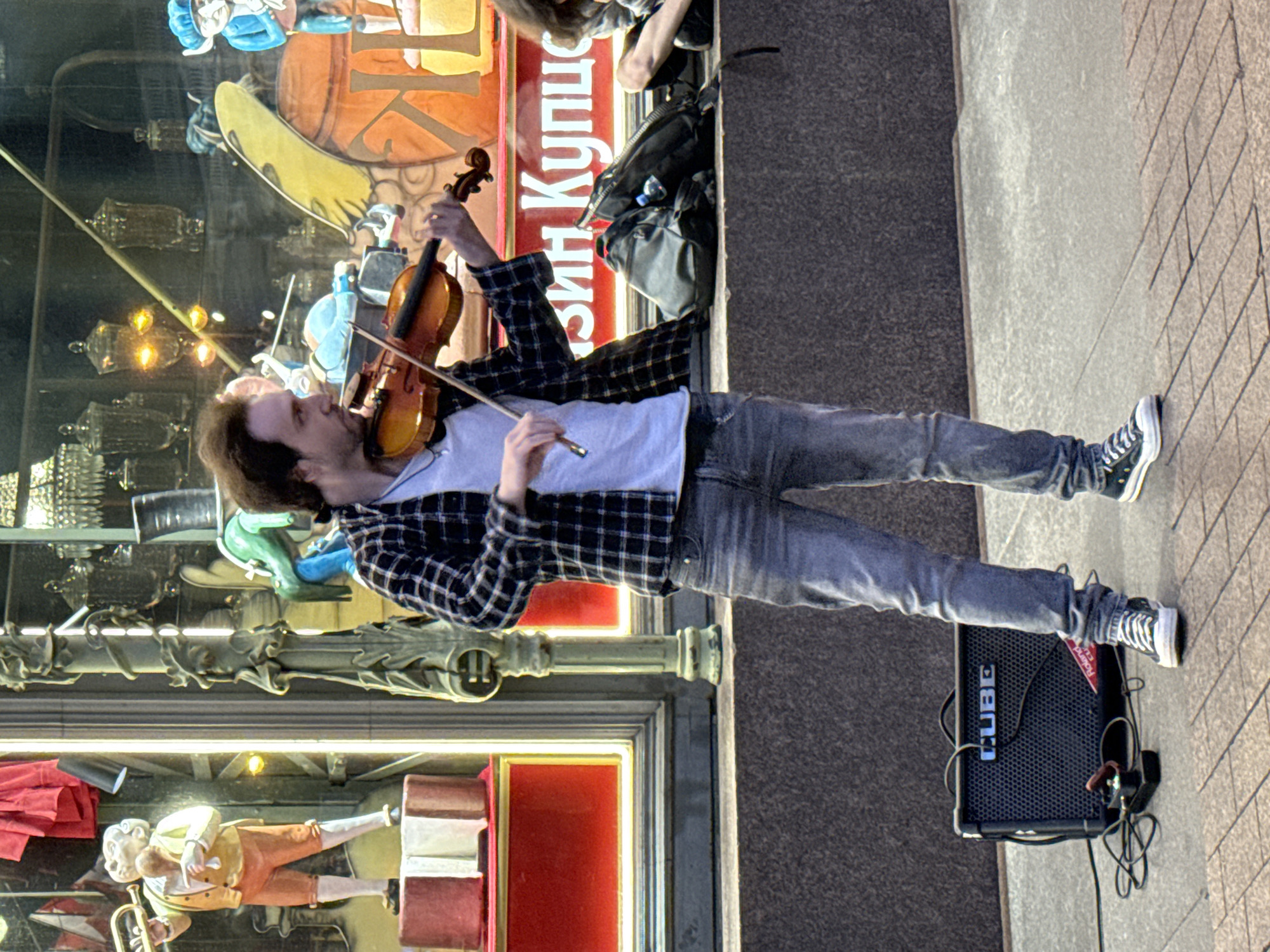 A man plays a violin on a city sidewalk in Saint Petersburg, Russia. He is dressed in a black and white plaid shirt, a white t-shirt, and blue jeans. He is holding a violin and bow, and appears to be performing. In front of him, on the ground, is a black amplifier with the word "CUBE" visible on it, and various wires and pedals. Behind him is a storefront window displaying various items, including figurines and decorative elements. The man is standing on a paved surface, with a textured wall behind him. The overall scene suggests a street performance in an urban environment.