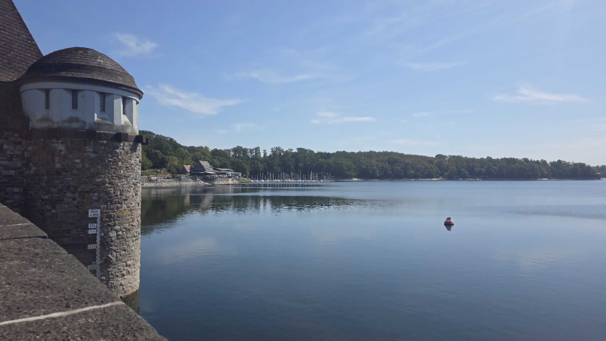 A tall, circular stone tower with a conical roof stands on the left side of the image, overlooking a calm body of water. The tower is constructed from rough, grey stones, and has small windows near its top. A dark, sloping roof covers the upper section of the tower. A water level marker with numbers is visible on the tower's stone facade. On the right side of the image, the water stretches to the horizon, reflecting the blue sky and scattered white clouds. In the middle of the water, a small, red and white buoy floats. Along the distant shore, a line of trees forms a dense green barrier, with a few buildings and a marina with numerous sailboats visible nestled among them. The overall scene depicts a serene landscape at the Möhnesee dam in Germany, under a clear, sunny sky.