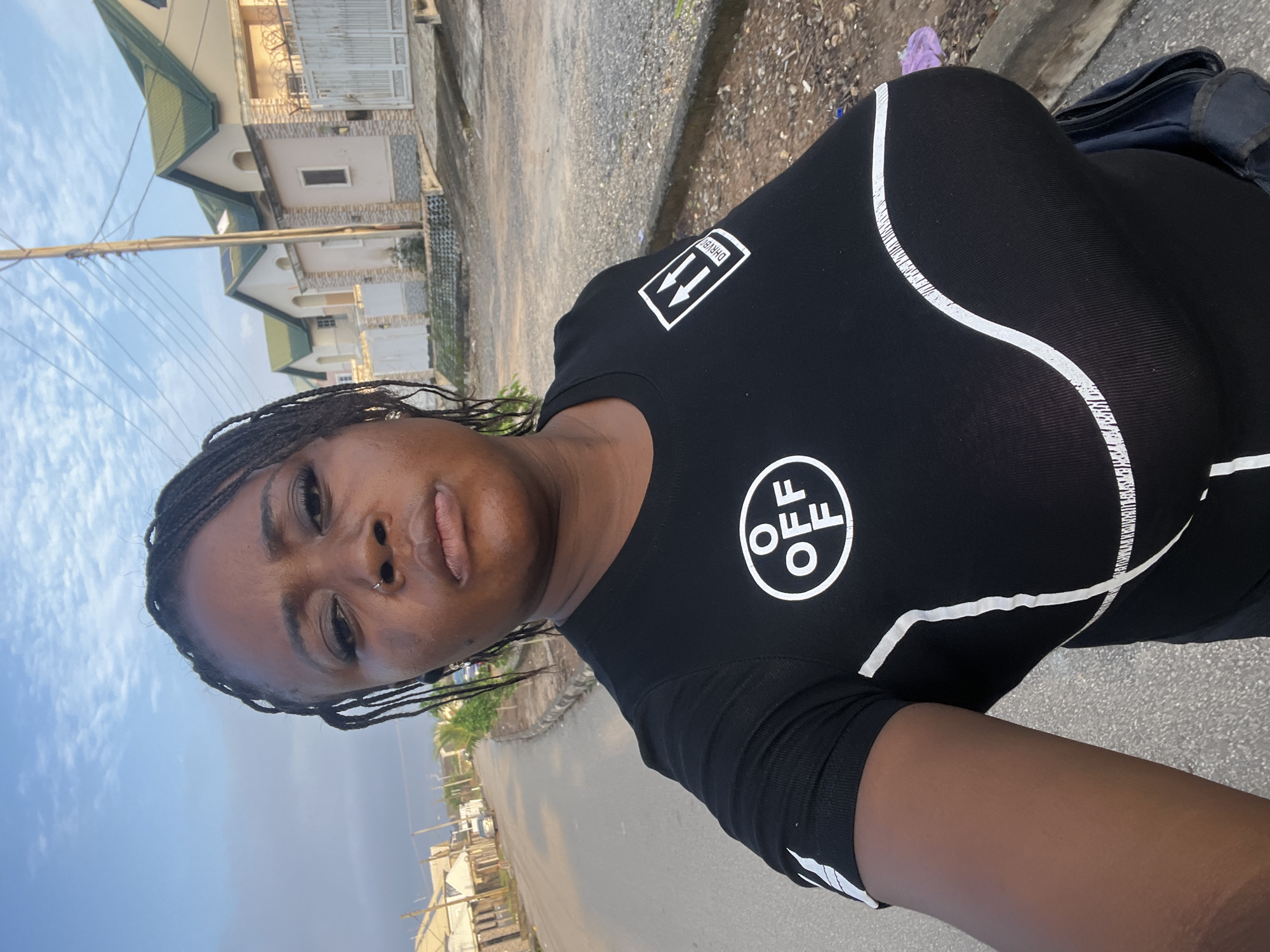 A woman with braided hair and a nose ring is standing on a paved street in Abuja, Nigeria. She is wearing a black t-shirt with white graphics. Behind her, there are modern houses with green roofs, a utility pole with wires, and a street with a few cars parked along the side. The sky is partly cloudy.