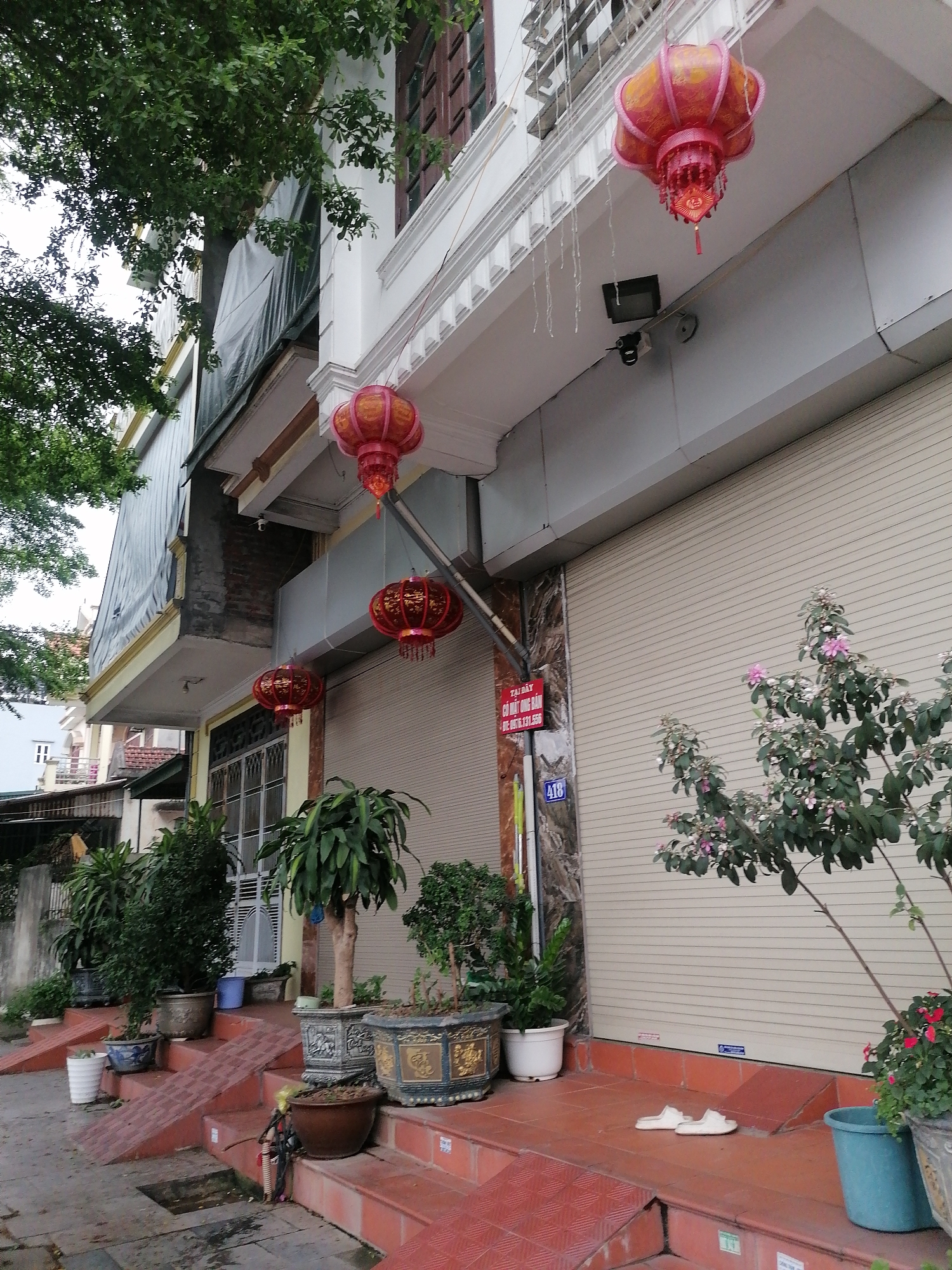 The scene captures the exterior of a building, situated in Ha Tu Ward, Vietnam. The building has a closed, beige-colored roll-up door. Above the door, a series of red and pink Chinese lanterns hang from the eaves. A security camera is mounted on the wall near the uppermost lantern. Plants in various pots line the base of the building and the steps leading up to the entrance. A sign in Vietnamese, mounted on the wall next to the roll-up door, is partially visible, with the number "418" on it. A pair of white slippers lies on the tiled landing in front of the closed door. The building is situated on a street with partial views of neighboring buildings and foliage from trees. The sky appears overcast.
