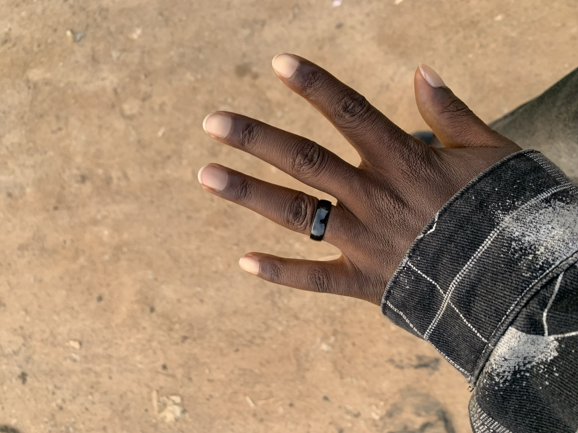 A close-up, overhead shot shows a dark-skinned hand with natural nails and a black ring on the ring finger. The hand is positioned over a sandy, dirt surface. The person is wearing a dark, checkered jacket with white stitching. The scene appears to be outdoors, possibly in a public area. The lighting suggests it's daytime.
