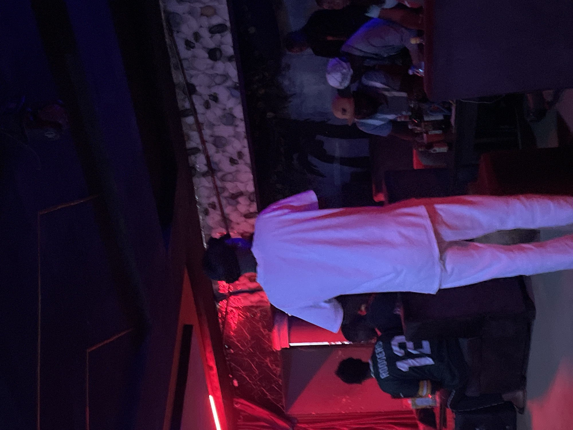 In a dimly lit venue in Durumi, Nigeria, several individuals are gathered. A person in the foreground is wearing a white t-shirt and pants, facing away from the camera. To the left, another person is seated, wearing a green and white jersey with the number "12" and the name "RODGERS" visible on the back. Behind them, several other people are seated around a table where bottles of drinks are present. The environment appears to be a bar or lounge, with red curtains and patterned walls. The lighting is low, with a mix of red and blue hues.