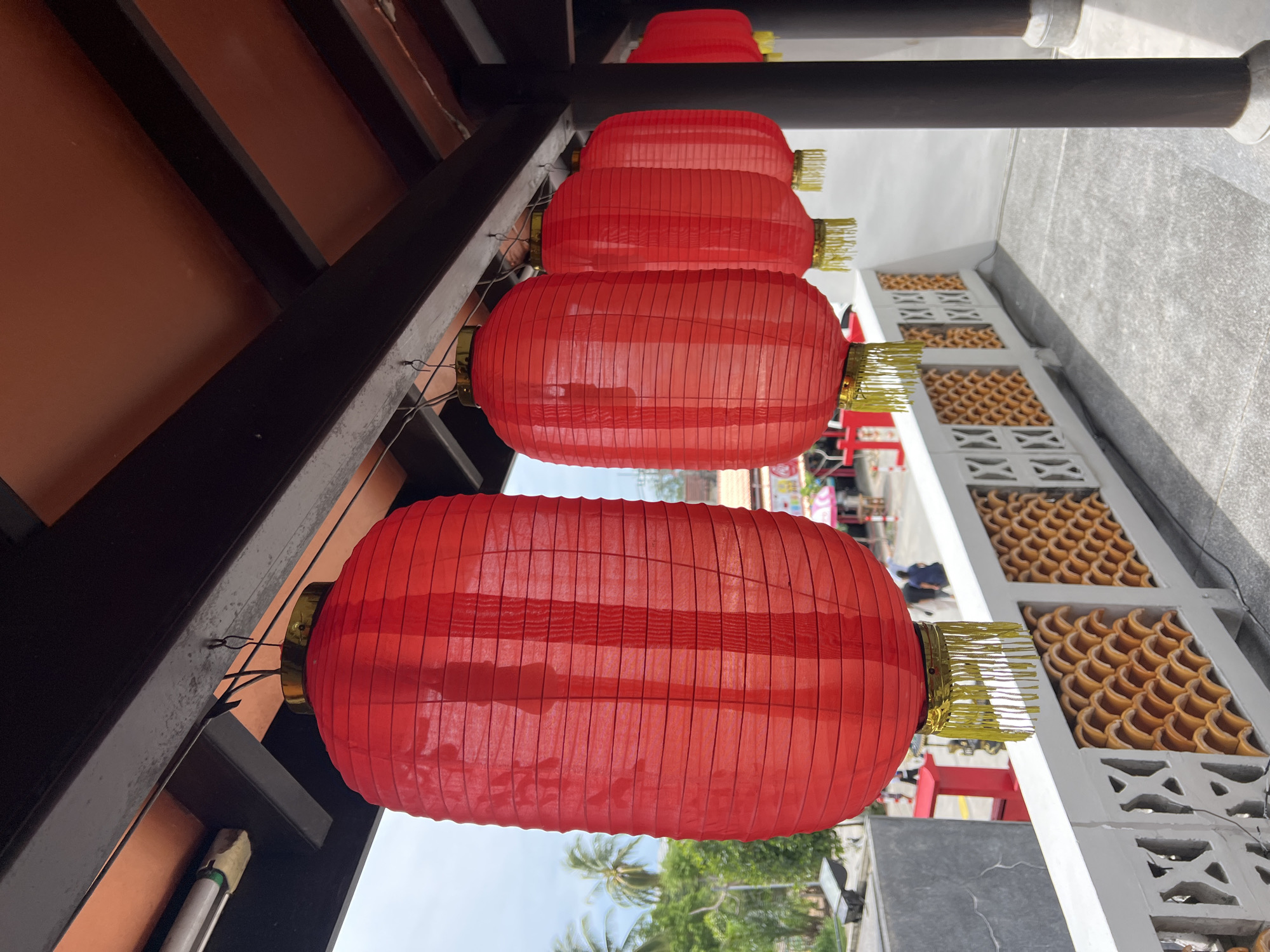 The image depicts a row of red, oval-shaped paper lanterns hanging from a dark brown, wooden overhang. Each lantern has a gold-colored top and is adorned with gold fringe tassels at the bottom. The lanterns are suspended by thin wires. In the background, a white railing with a patterned design is visible, behind which a street scene unfolds with palm trees, buildings, and a few people. The location is identified as Baan Hua Thanon, Thailand. The overall scene suggests a festive or cultural setting, possibly a market or a temple area, under a bright, overcast sky.