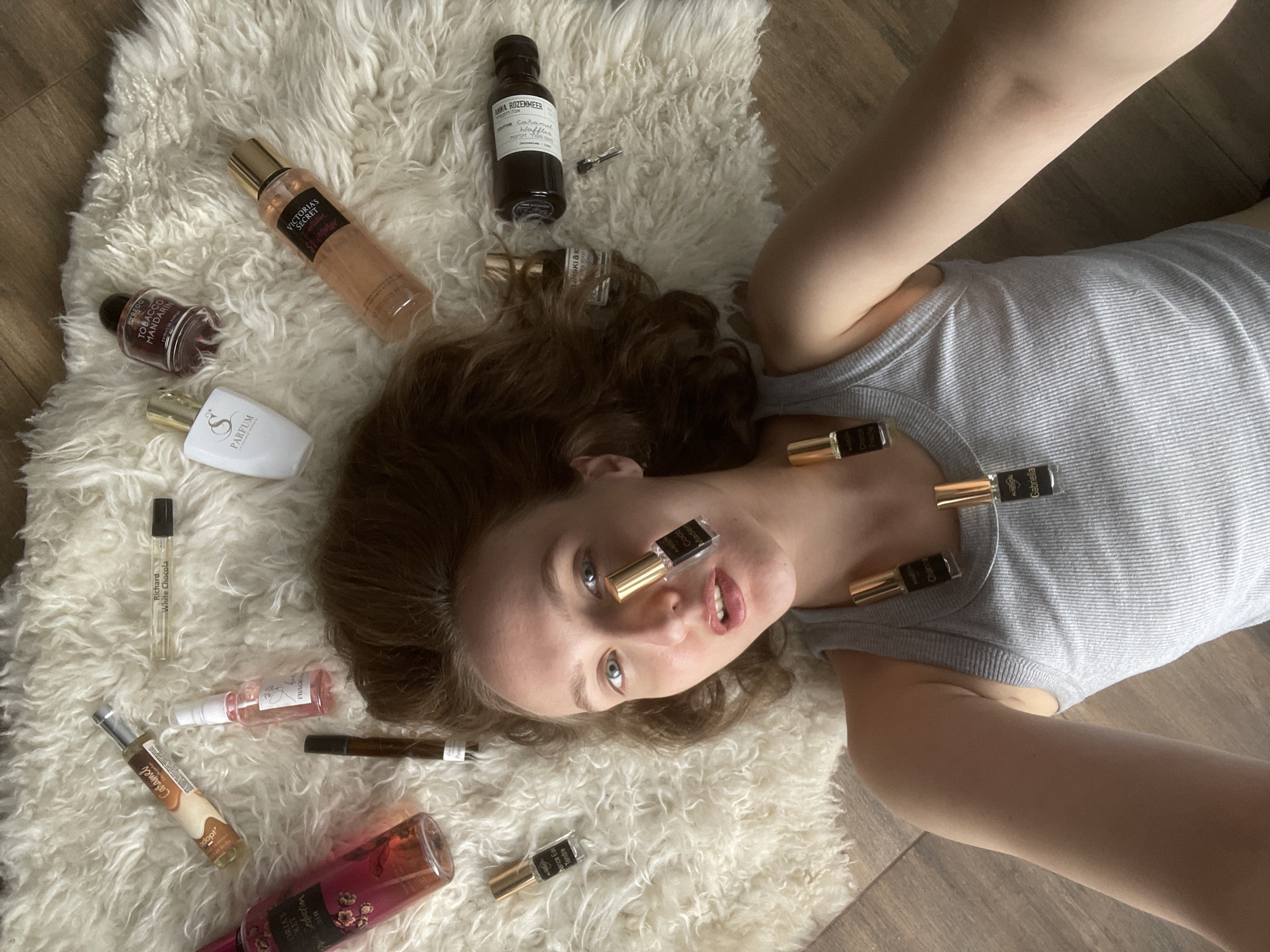 A young woman is lying on a white, shaggy rug on a wooden floor. She is wearing a gray tank top and is surrounded by various perfume bottles. Some of the bottles are scattered on the rug, while others are positioned on her face and neck. The bottles vary in size and shape, with some being full-sized and others being smaller samples. The room appears to be indoors and well-lit. There is no mention of the location in the city of Подшивалово, Russia.