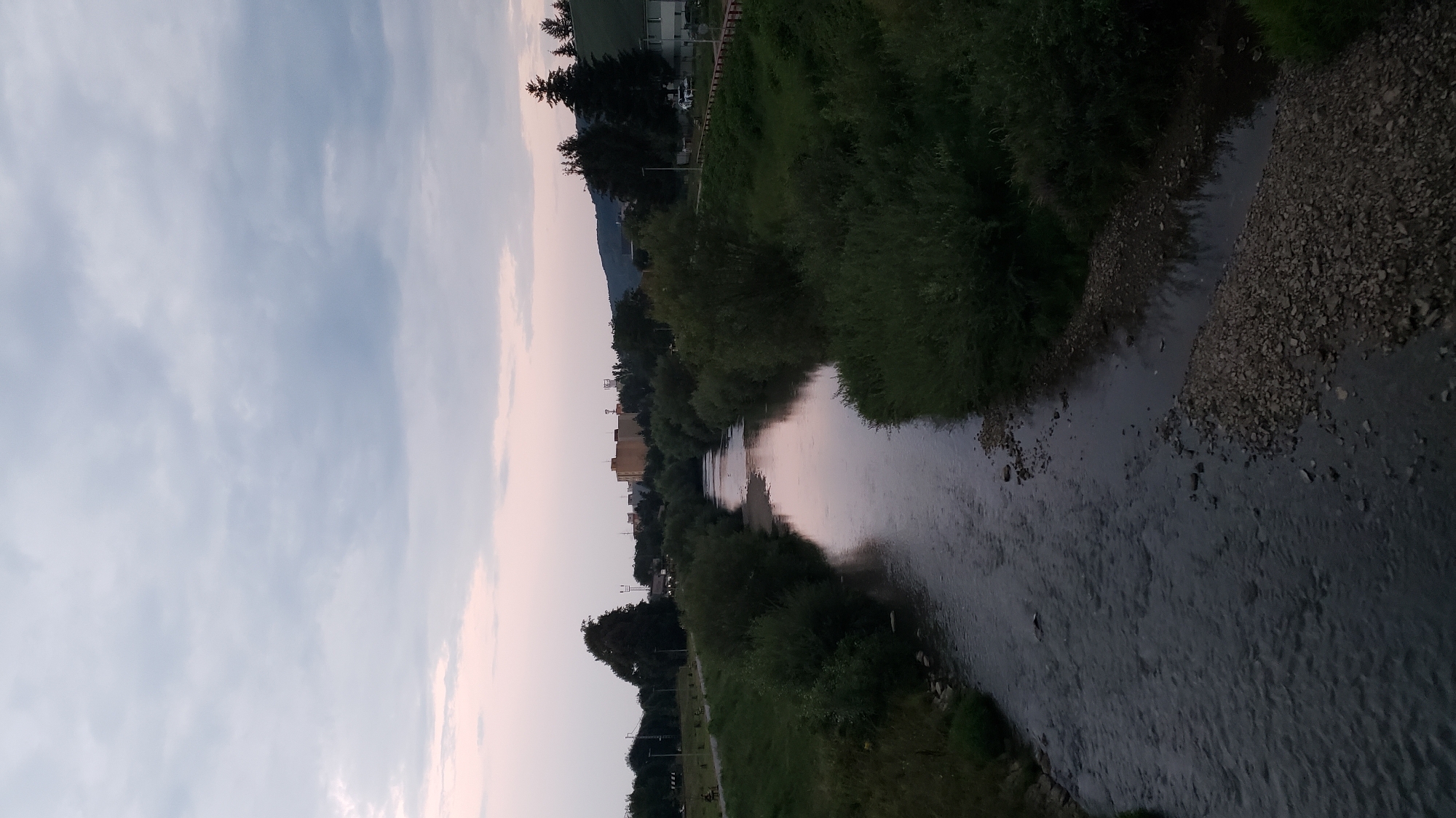 This is a photograph taken from a bridge, looking down at a river flowing through a city. The river is surrounded by lush green trees and bushes. In the distance, there are several buildings, including some taller structures that appear to be industrial or commercial. The sky is overcast with gray clouds, and there is a hint of pink and purple light in the sky, suggesting it is either dawn or dusk. The water in the river is dark and reflective, with some ripples on its surface. The riverbanks are lined with rocks and vegetation. There are no people visible in the image, nor any specific actions or interactions taking place. The scene depicts a natural landscape within an urban environment. This view is likely from a bridge over the Torysa River in the city of Prešov, Slovakia, given the presence of urban infrastructure juxtaposed with the river and greenery.