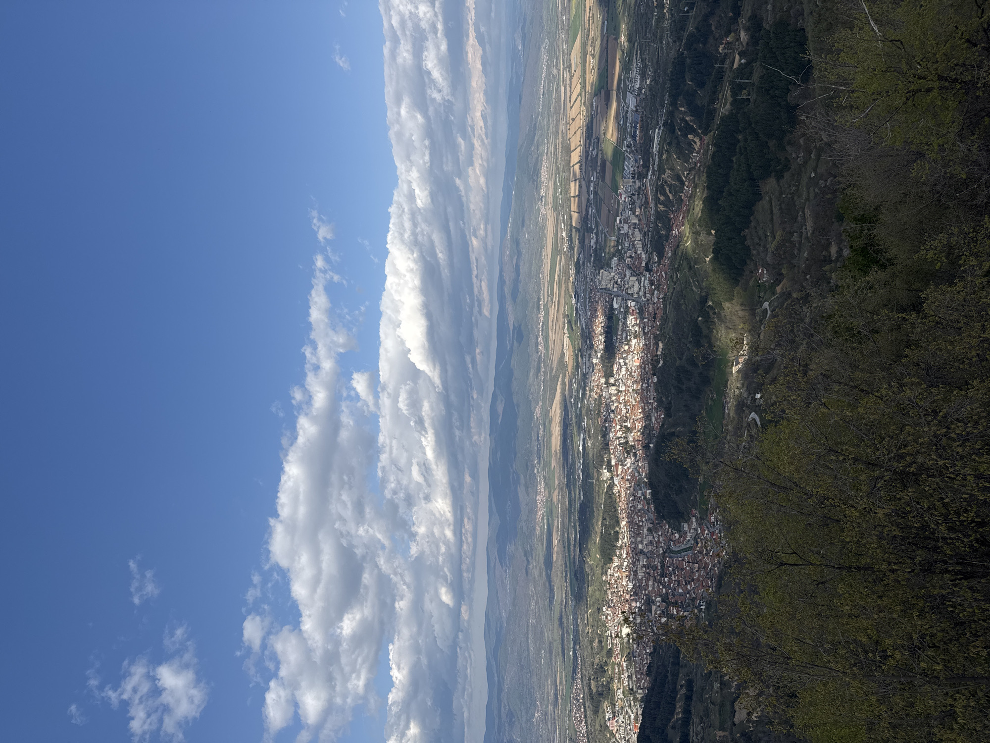The image presents a high-angle, aerial view of a landscape featuring a town nestled in a valley. On the left side of the frame, a blue sky is visible with scattered cumulus clouds. To the right, the town's buildings are tightly packed, showing a mix of white and red roofs. The valley floor contains agricultural fields of varying colors, indicating different crops or stages of growth. The topography is hilly, with forested areas and rocky terrains visible. In the foreground, foliage from trees or bushes is seen, partially obscuring the view. The town is likely Delchevo, Bulgaria.