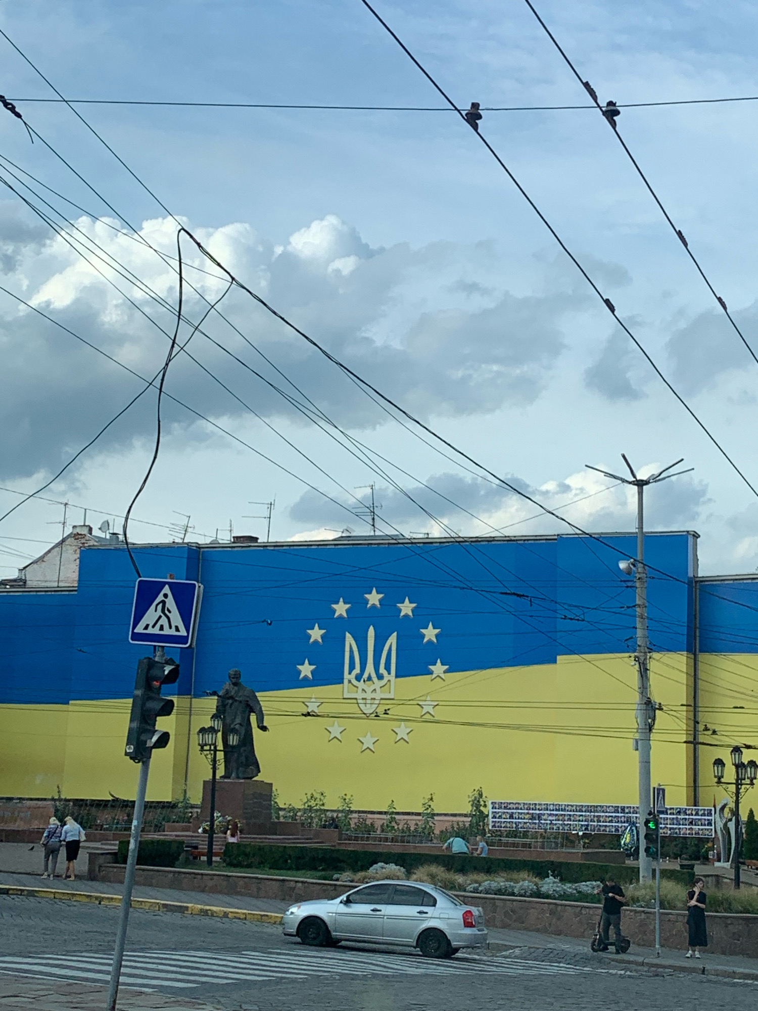 The image shows a street scene in Chernivtsi, Ukraine, with a large building painted with the Ukrainian flag colors and symbols. A statue of a man stands in front of the building. In the foreground, there are traffic lights, a pedestrian crossing, and a silver car. Several people are visible walking or standing on the sidewalk. Overhead, numerous electrical wires crisscross the sky. The sky is cloudy.