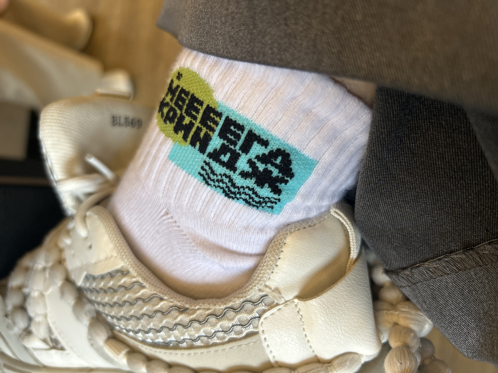 The image shows a close-up of a person's foot wearing a white sock with a design that includes green and blue elements, along with text that reads "HEEERA KPNHAжK." The person is wearing an off-white sneaker with a textured design and a visible tag that says "BL869." The foot is positioned near a piece of grey denim fabric. The location, Plovdiv, Bulgaria, cannot be confirmed based solely on the visual information present in the image. There are no identifiable landmarks or indicators to determine a specific location.