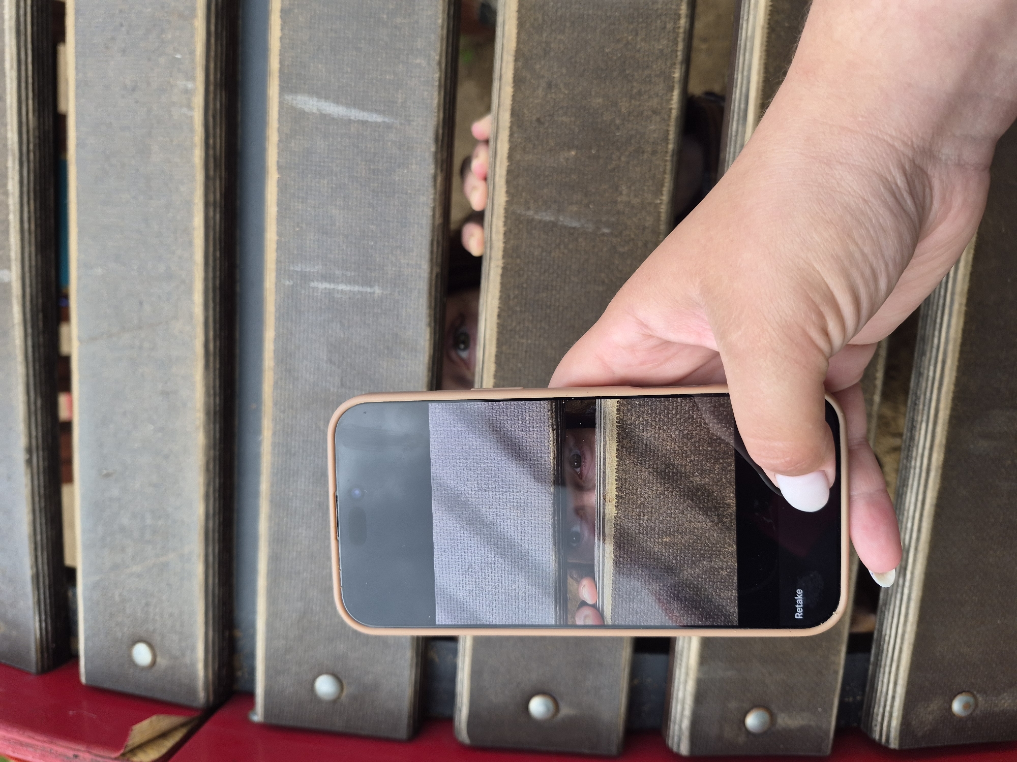 A person is holding a smartphone in their right hand, taking a photo of a second person peeking through narrow vertical wooden slats. The slats are gray with brown edges and secured with round, silver-colored fasteners. The smartphone screen displays a close-up of the peeking person's eyes. The scene takes place on what appears to be a red bench-like structure.