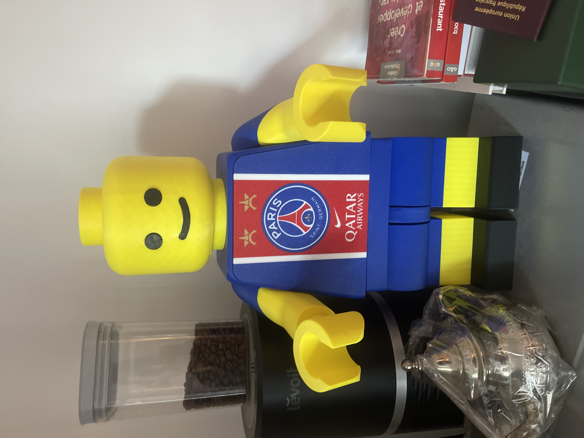 The image shows a large, yellow Lego-style figure dressed in a blue and red Paris Saint-Germain football club jersey with yellow accents. The figure is standing in front of a white wall. To the left of the figure is a clear plastic container filled with coffee beans, sitting on top of a black electric device with "Lĕvoit" branding. In the foreground to the left, partially covered in plastic wrap, is a metallic teapot. To the right of the figure, a stack of books is visible. The setting does not provide any specific indicators to pinpoint a location within the city of Paris, France. The scene depicts static objects and a figurine, with no people or actions.