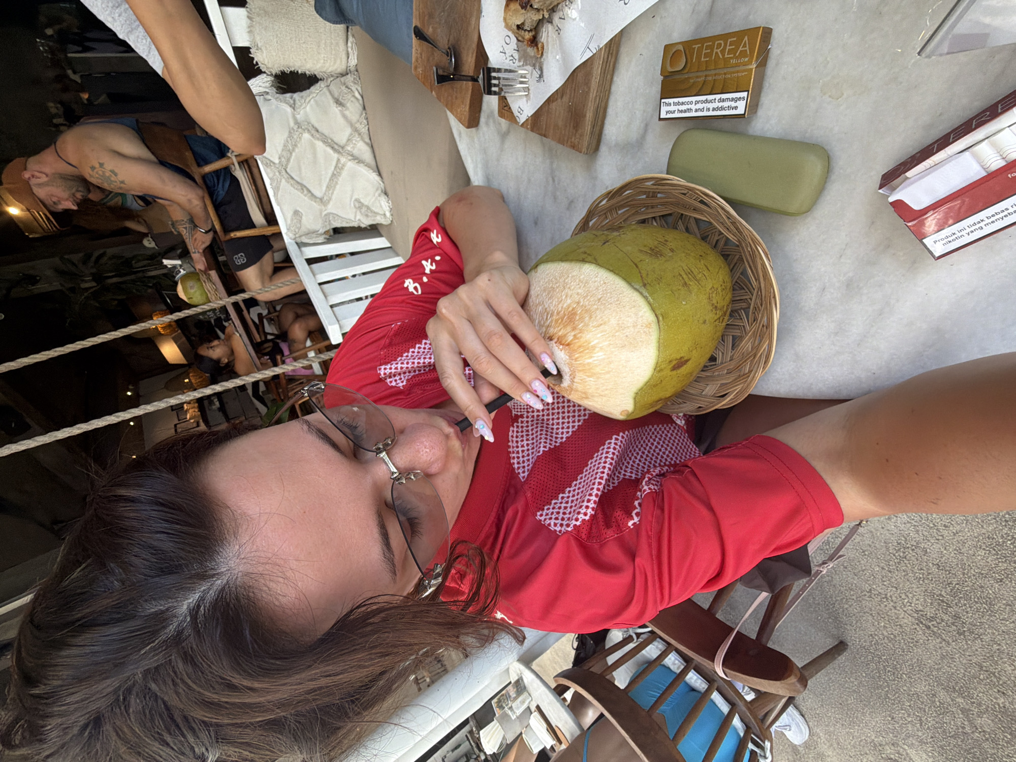 In North Kuta, Indonesia, a woman is seated at a marble table, holding a straw to a halved young coconut with a woven placemat. She is wearing a red shirt with white diagonal stripes and is looking down at the coconut. A wooden chair with blue cushions is visible to her left. In the background, a man wearing a gold hat and a dark blue tank top is seated at another table, facing away from the camera. Another person's arm, wearing a grey sleeve, is visible in the upper right corner. A swing seat with white cushions is hanging in the background. On the table, in addition to the coconut, are two wooden cutting boards, one with a fork and spoon, and another with a pastry. A green case and two cigarette packs are also on the table. The scene appears to be an outdoor dining area.