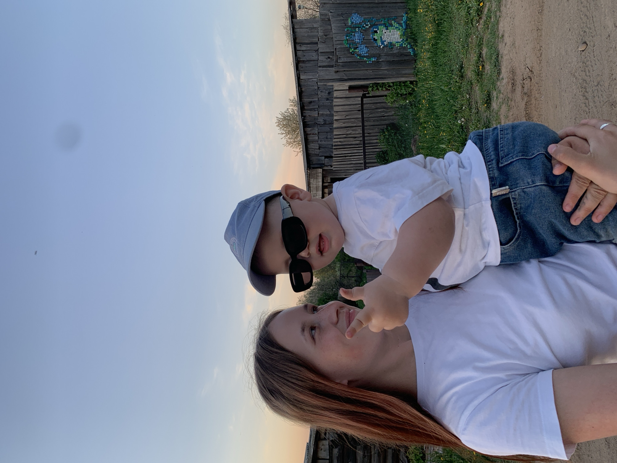 A child wearing sunglasses and a blue cap is sitting on a woman's chest. Both are wearing white t-shirts. The child is pointing forward. The woman is looking up at the child. They are outside in front of a wooden building decorated with an art piece made from colorful bottle caps. The location is Фоки, Russia. The sky is visible with blue transitioning to a lighter hue near the horizon. The ground is visible with dirt and some patches of grass.