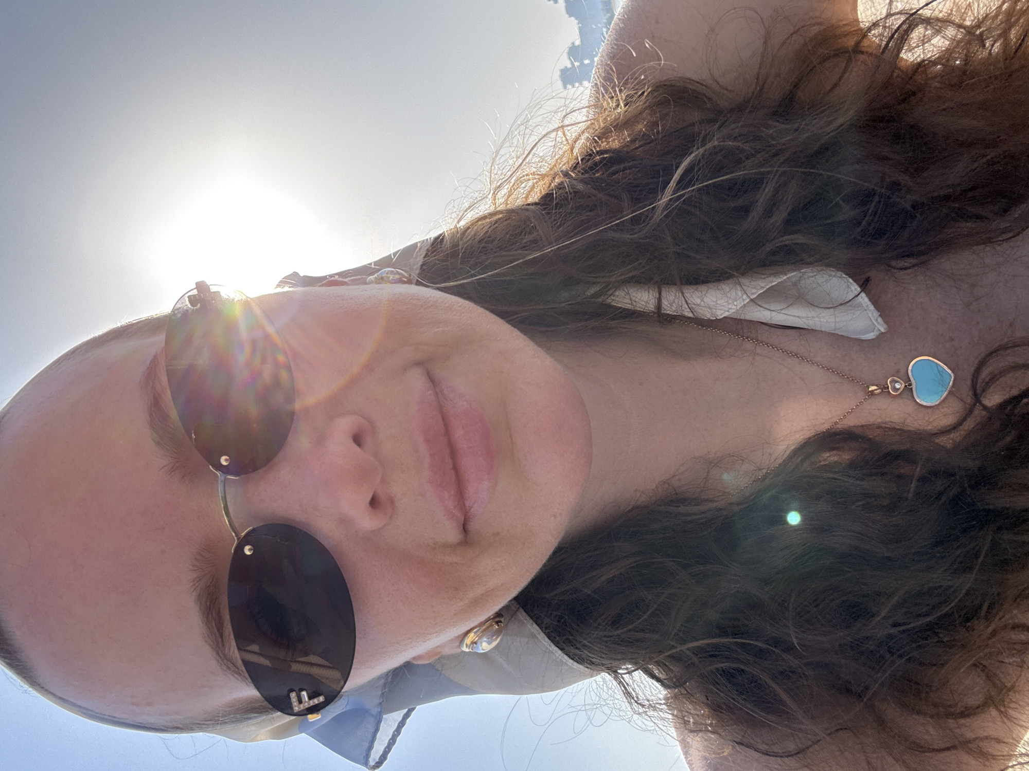 A woman is captured in a close-up selfie, positioned in Dubai, United Arab Emirates. She is wearing oval-shaped, dark sunglasses with gold frames, adorned with a gold "F" emblem and small diamonds on the left lens. A light-colored headscarf, possibly silk, is tied around her head, with a portion visible on the left side of the frame. She has long, wavy brown hair. She is wearing gold hoop earrings and a rose gold necklace with a heart-shaped blue pendant and a small diamond. The bright sun is shining from the upper right, causing lens flare and overexposure in the background, which appears to be a clear sky. In the far background, some blurred structures are visible. The woman's expression is neutral as she looks directly at the camera.