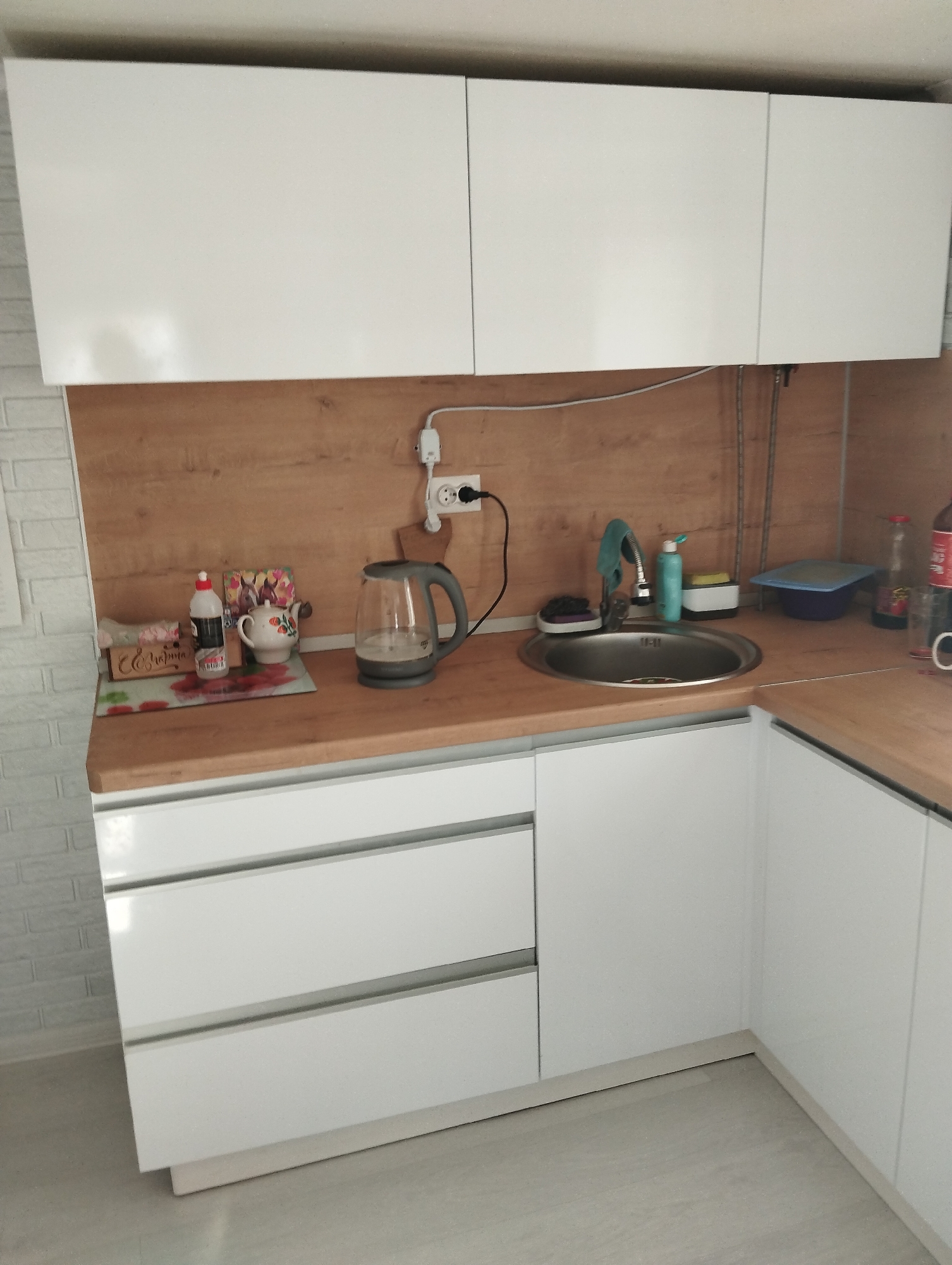 A kitchen scene featuring white cabinets and drawers, a wooden countertop with a sink, a kettle, cleaning supplies, and food items. The backsplash is also wooden. A power outlet with a plugged-in kettle is visible. The location is Krasnogorskoye, Russia.