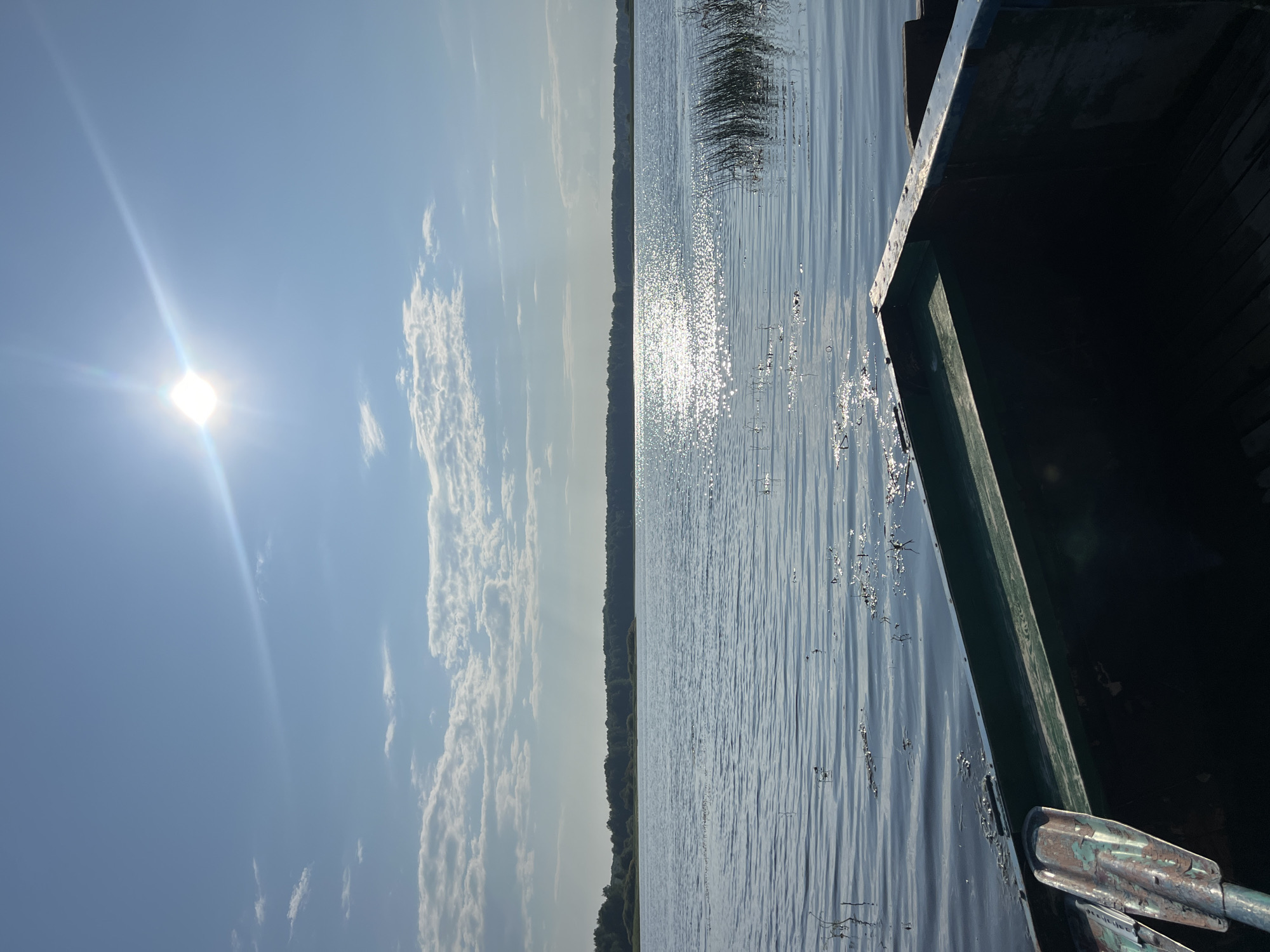 The image depicts a serene lake scene under a bright, sunny sky with scattered clouds. The sun is positioned high in the sky, creating a brilliant reflection on the water's surface. In the foreground, the interior of a dark-colored rowboat is visible, with an oar lying inside. The lake stretches out to a tree-lined horizon, suggesting a natural, possibly rural setting. There are no people visible in the image. The environment is calm and tranquil. Based on the provided information, this scene is located in the city of Kondas, Russia.