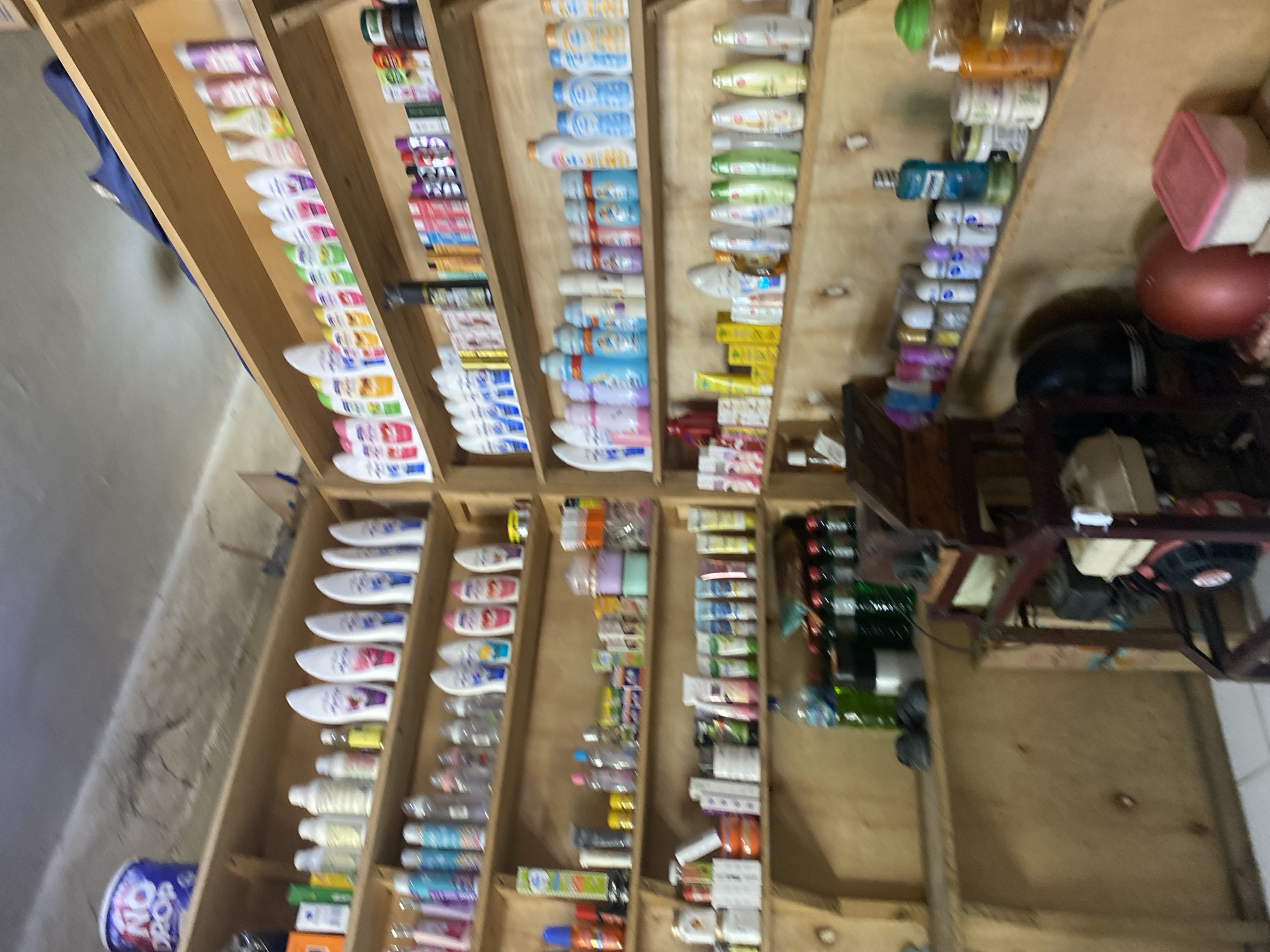The image depicts a retail shelf display filled with various personal care products and household items, suggesting a small shop or market stall. The shelves are made of unfinished wood and are packed with an assortment of bottles, tubes, and containers. Products include shampoos, lotions, soaps, and possibly cleaning supplies. Some items are organized by color or type, while others appear to be more randomly placed. The background shows a rough, possibly concrete or stucco wall, and a portion of a corrugated metal ceiling. The lighting appears to be artificial, casting a warm glow on the products. There is a piece of machinery or equipment on a lower shelf towards the right side of the image, which could be related to the operation of the shop. Based on the context of personal care products and the typical retail environment, this scene is likely located in a commercial area of Ta', Indonesia.
