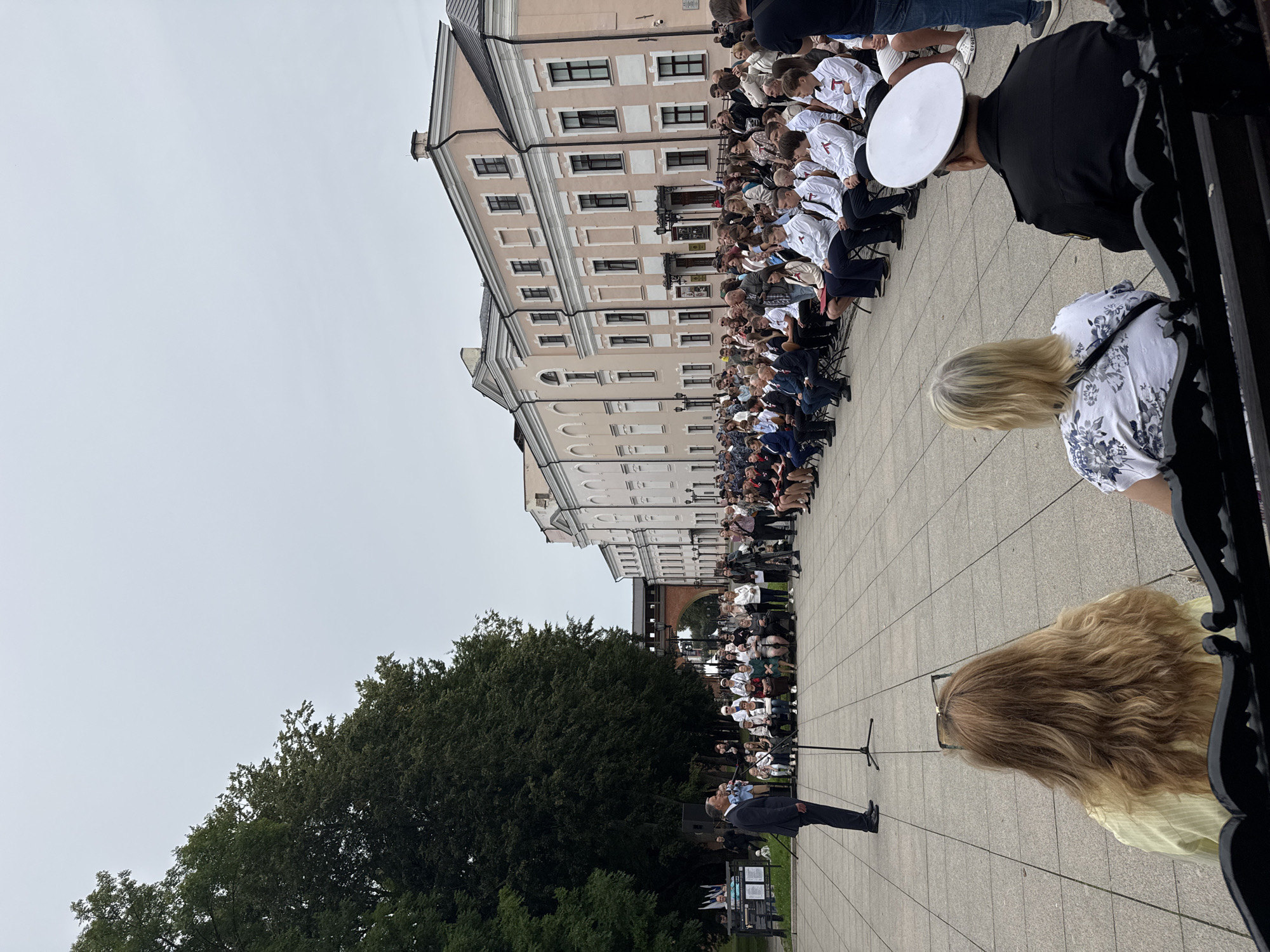 An outdoor gathering is taking place on a paved square in Veliky Novgorod, Russia, in front of a pale pink, multi-story building with classical architectural elements. A man in a dark suit is standing at a podium and addressing a crowd of people seated in rows. Many of the seated individuals, particularly those in the front rows, appear to be young men and women wearing white shirts with red accents, possibly a uniform or ceremonial attire. A large tree with dense foliage is visible to the left of the scene, and the sky is overcast. In the foreground, two women are seated on a bench, facing away from the camera, observing the event. To the right, a man in a dark uniform with a white naval-style hat is also seated on a bench. The overall atmosphere suggests a formal event or ceremony.