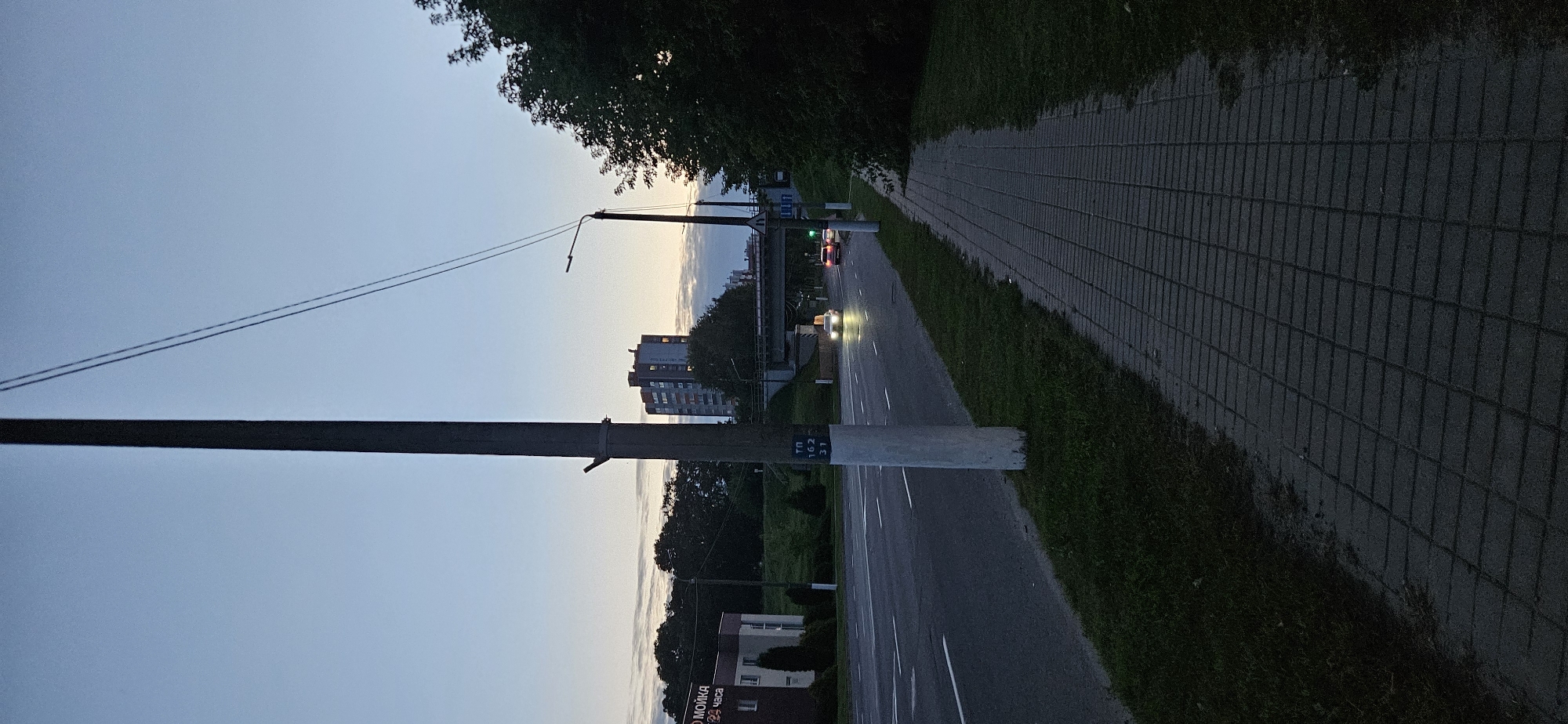 The image depicts a street scene at dusk in Vitsebsk, Belarus, likely in the evening due to the dim lighting and headlamps of approaching vehicles. A concrete sidewalk, paved with rectangular bricks, runs along the right side of the frame, bordered by a strip of grass and then a two-lane road. Two cars, one white and one red, are visible on the road, their headlights illuminated. In the background, a multi-story building with illuminated windows stands against a twilight sky. A pedestrian overpass crosses the road in the mid-ground. A tall, dark utility pole, likely for electrical or telecommunications lines, dominates the left side of the foreground, with wires extending from it. Another similar pole with a streetlamp is visible further down the road on the right. A small, dark-colored building with signage in Russian is present on the far left of the scene. The overall environment suggests an urban setting during the transition from day to night.