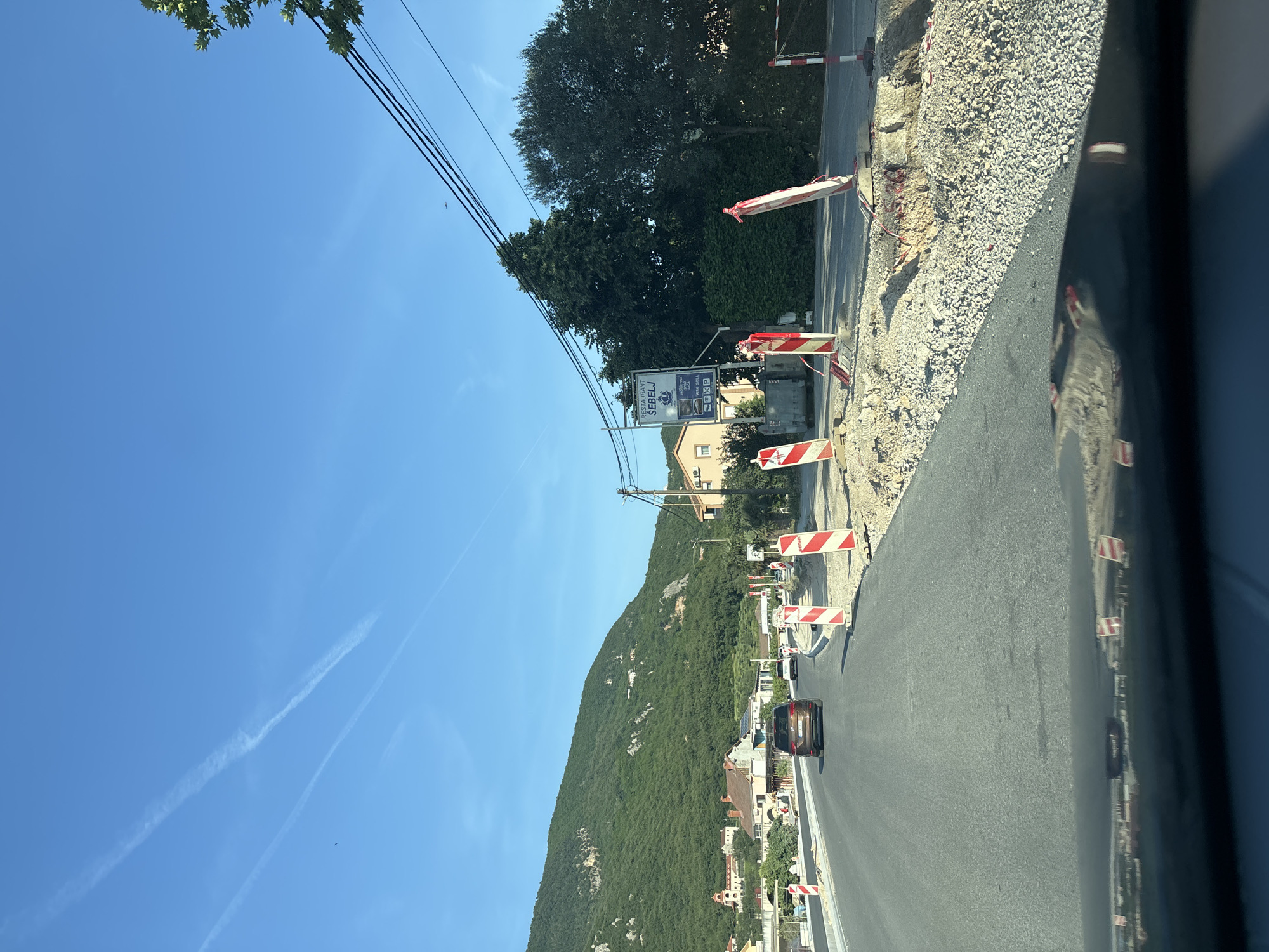 The image depicts a bright, sunny daytime scene of a road in a mountainous coastal region, likely captured from inside a moving vehicle. The perspective is angled downwards, showing the asphalt road in the foreground and a brown car ahead.
To the right of the road, there's an active construction or renovation area, characterized by a freshly laid gravel shoulder and loose earth. Red and white striped construction barriers are placed along the edge of this area, separating it from the paved road. A noticeable reflection of the road, sky, and possibly parts of the interior of the vehicle is visible on the far right, suggesting a window.
On the left side, a verdant, tree-covered mountain rises prominently under a clear blue sky. Scattered houses and buildings with typical Mediterranean architecture are nestled at the base of the mountain and along the road. Power lines run across the upper part of the image, partially obscured by tree foliage.
A large sign for "RESTAURANT SEBELJ" is visible on the left, near the road. Below the restaurant name, icons and text advertise its offerings, including "RIBLJI SPECIJALITETI" (Fish Specialties), "MESO SA ŽARA" (Grilled Meat), and indicate amenities like Wi-Fi and parking ("P").
The sky is a brilliant clear blue, with several distinct white contrails streaking across it. The overall impression is one of travel through an area undergoing development, surrounded by natural beauty.