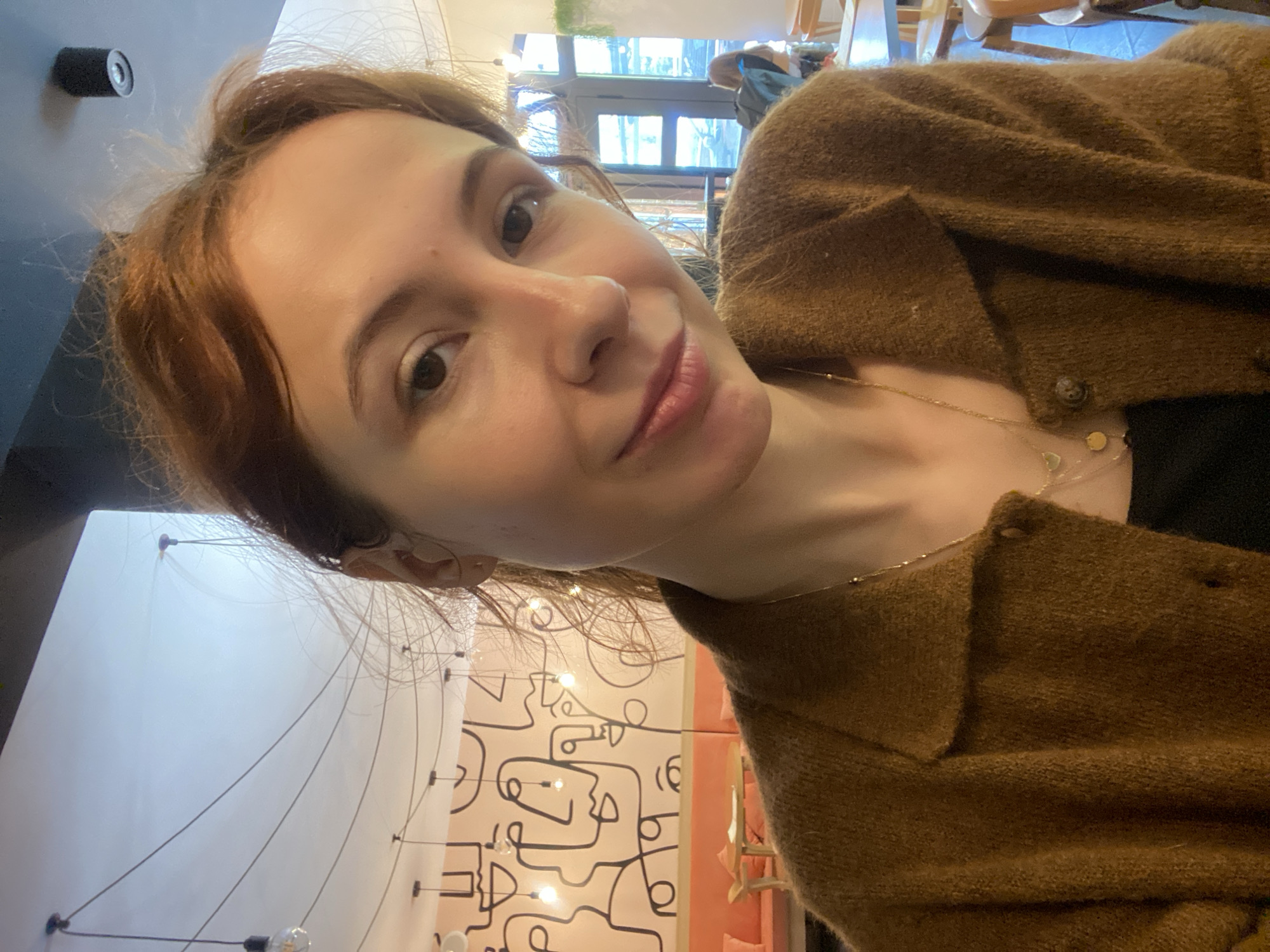 A woman with reddish-brown hair, wearing a brown cardigan and a gold necklace, is taking a selfie. The background shows a light-colored wall with abstract black line art and a lighting fixture with dangling bulbs. A window is visible in the upper part of the frame, along with glimpses of the interior of the building. Based on the art style and interior design, the location is likely a cafe or restaurant in Novi Sad, Serbia, known for its modern and artistic ambiance.