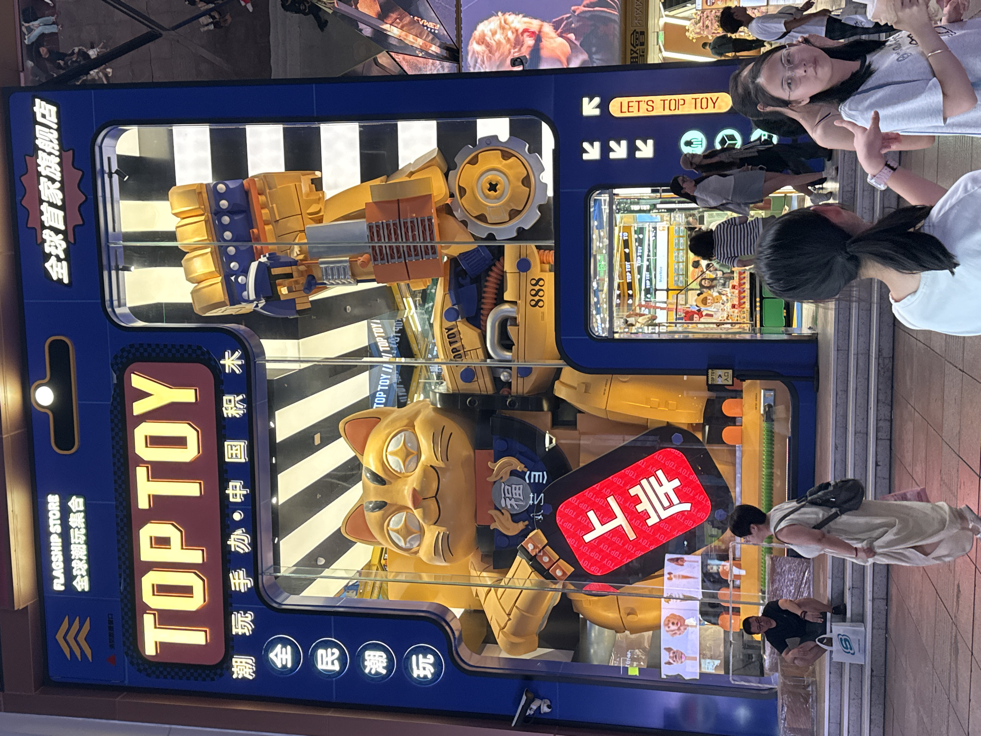 An external view captures a brightly lit storefront in Shanghai, China, featuring a large, yellow, robotic cat mascot. The storefront itself is designed to resemble a toy packaging box, with "TOP TOY" prominently displayed in illuminated lettering. The mascot is a prominent display, adorned with a red tag bearing white Chinese characters that read "上岸" (meaning "to land" or "to make a breakthrough"). The store appears to be a flagship branch, indicated by the Chinese text "全球首家旗舰店" (Global First Flagship Store) above the entrance.
Several individuals are visible in the scene. A man in a black t-shirt and shorts is seated on the steps in front of the store, with a white shopping bag at his feet. A woman in a beige dress with a backpack walks away from the store. Inside the store, through the glass, several people can be seen browsing. Further out in front of the store, a group of young women are standing and walking, with one in the foreground looking towards the camera and holding a drink. The environment suggests a commercial street or shopping district at night, with reflections of lights and other buildings visible on the storefront glass. The overall impression is of a vibrant and modern commercial setting.