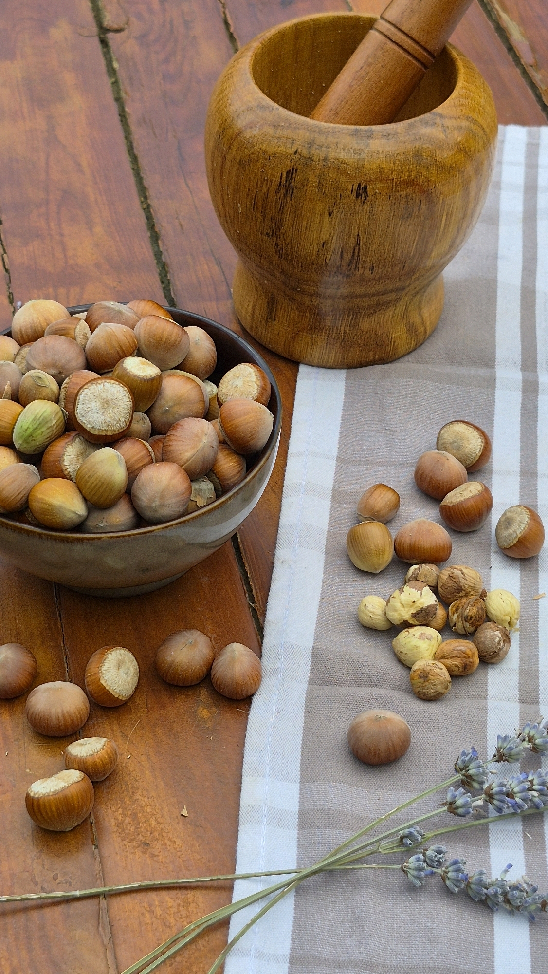 A close-up, overhead shot captures a rustic arrangement of hazelnuts and a wooden mortar and pestle on a weathered wooden surface. A beige ceramic bowl, filled with hazelnuts, sits to the left of the frame. The hazelnuts are a mix of whole nuts with shells and some cracked open, revealing their pale interiors. To the right of the bowl, a striped beige and white cloth is laid out, with more hazelnuts scattered on top. Some of these hazelnuts are shelled, showing their lighter color. A wooden mortar and pestle, with a smooth, polished finish, is positioned in the upper right corner, the pestle resting inside the mortar. In the lower right corner, a few sprigs of dried lavender are arranged on the cloth, adding a subtle touch of color and fragrance to the scene. The lighting is natural and diffused, creating soft shadows and highlighting the textures of the wood and nuts. The overall impression is one of natural simplicity and preparation for a culinary task.