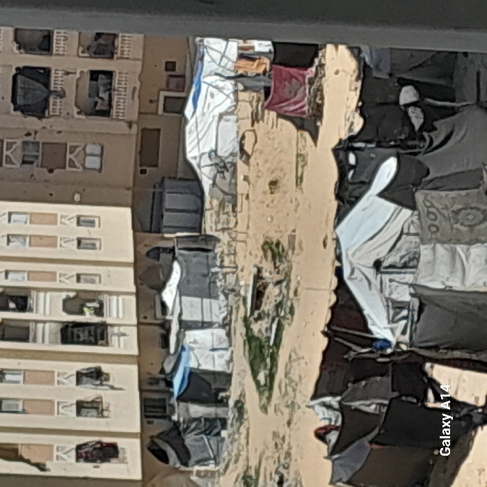 The image depicts a crowded informal settlement in Khan Yunis, Palestinian Territory, viewed from an elevated perspective. The scene is dominated by numerous densely packed, makeshift shelters constructed from tarpaulins, dark fabrics, and blankets, scattered across sandy, uneven ground with sparse vegetation. In the foreground and midground, clusters of these shelters, varying in color from dark greys and blacks to whites and light blues, are visible. Some fabric items are draped or hung around them. In the background, multi-story residential buildings with beige facades and numerous windows, some appearing weathered or damaged, rise above the settlement. Additional shelters are constructed at the base of these buildings. No identifiable individuals or specific actions are clearly discernible within the frame. The "Galaxy A14" watermark is present in the bottom-left corner.