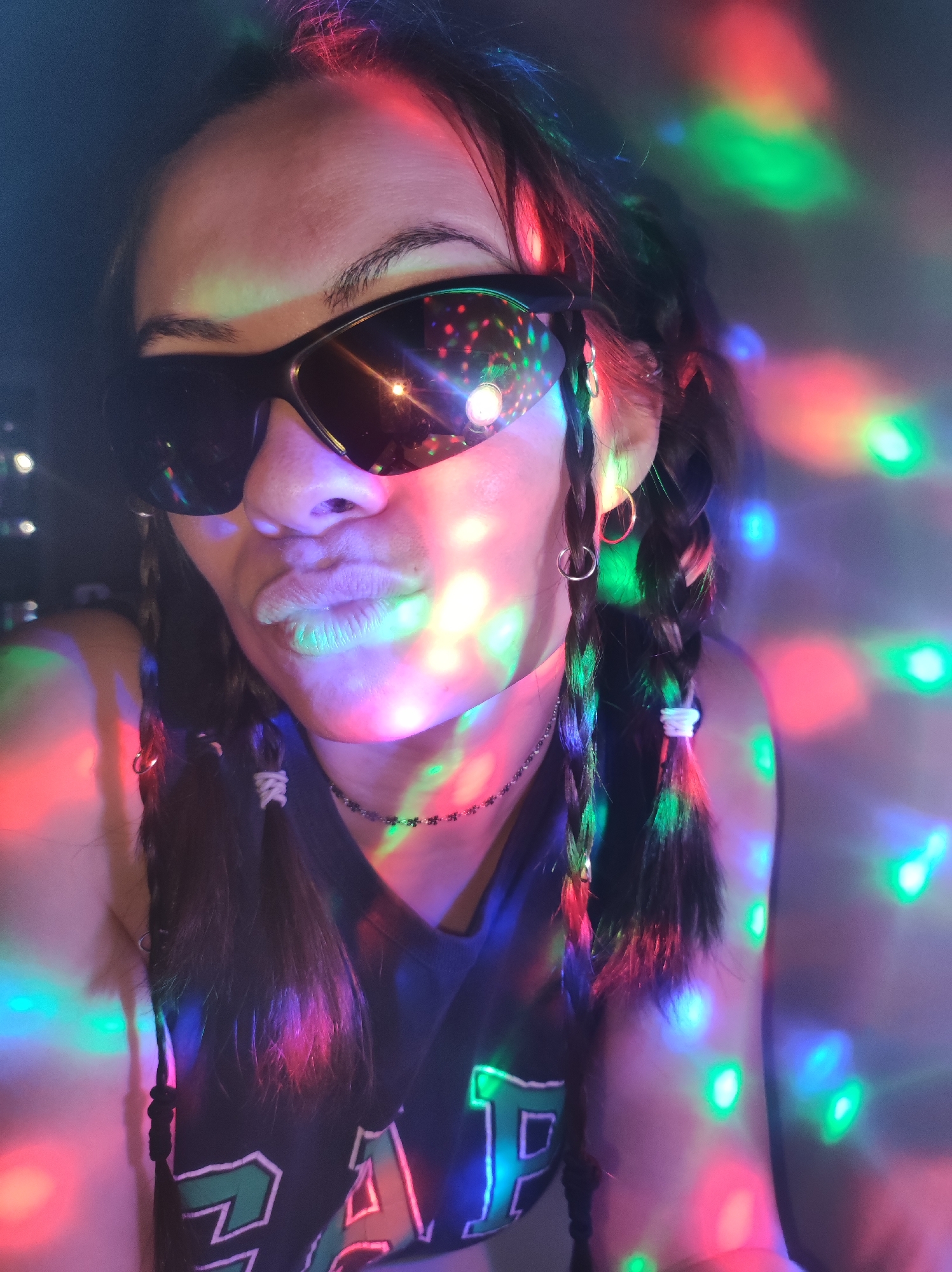 A person is captured in a close-up, chest-up portrait, adorned with dark sunglasses and striking a pouting pose with their lips. Their dark hair is styled in multiple braids, some secured with white elastic bands and others featuring small metallic rings. Large hoop earrings are visible, and a choker necklace with small flower or star-shaped links graces their neck. The individual wears a dark tank top or shirt, upon which green-outlined letters, possibly "P" and "A" from a larger word, are visible on the front.
The scene is dramatically lit by numerous circular, multi-colored light reflections (red, green, blue), characteristic of a disco ball or party lights. These vibrant spots scatter across the person's face, hair, and clothing, as well as the dark, blurred background, creating a lively, nocturnal, or indoor party atmosphere. Bright reflections are particularly noticeable on the sunglasses, showing miniature versions of the colored lights. The overall mood conveyed is playful and energetic. The image was taken in Фоки, Russia.