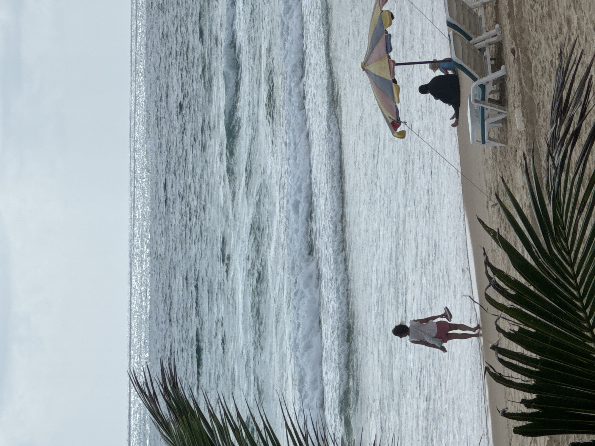 The image depicts a scenic view of Karon Beach in Karon, Thailand, under an overcast sky. In the foreground, lush green palm fronds partially obscure the sandy beach. Several lounge chairs are arranged on the sand, with one occupied by a person wearing a black headscarf and a child in a blue and red swimsuit sitting beside them under a colorful, multi-striped umbrella. Further down the beach, a lone woman walks towards the water, carrying a pair of flip-flops in her hand. She is wearing a white top and pink shorts. The ocean displays moderate waves, with the water shimmering under the diffused sunlight. The horizon is clearly defined where the sea meets the sky.