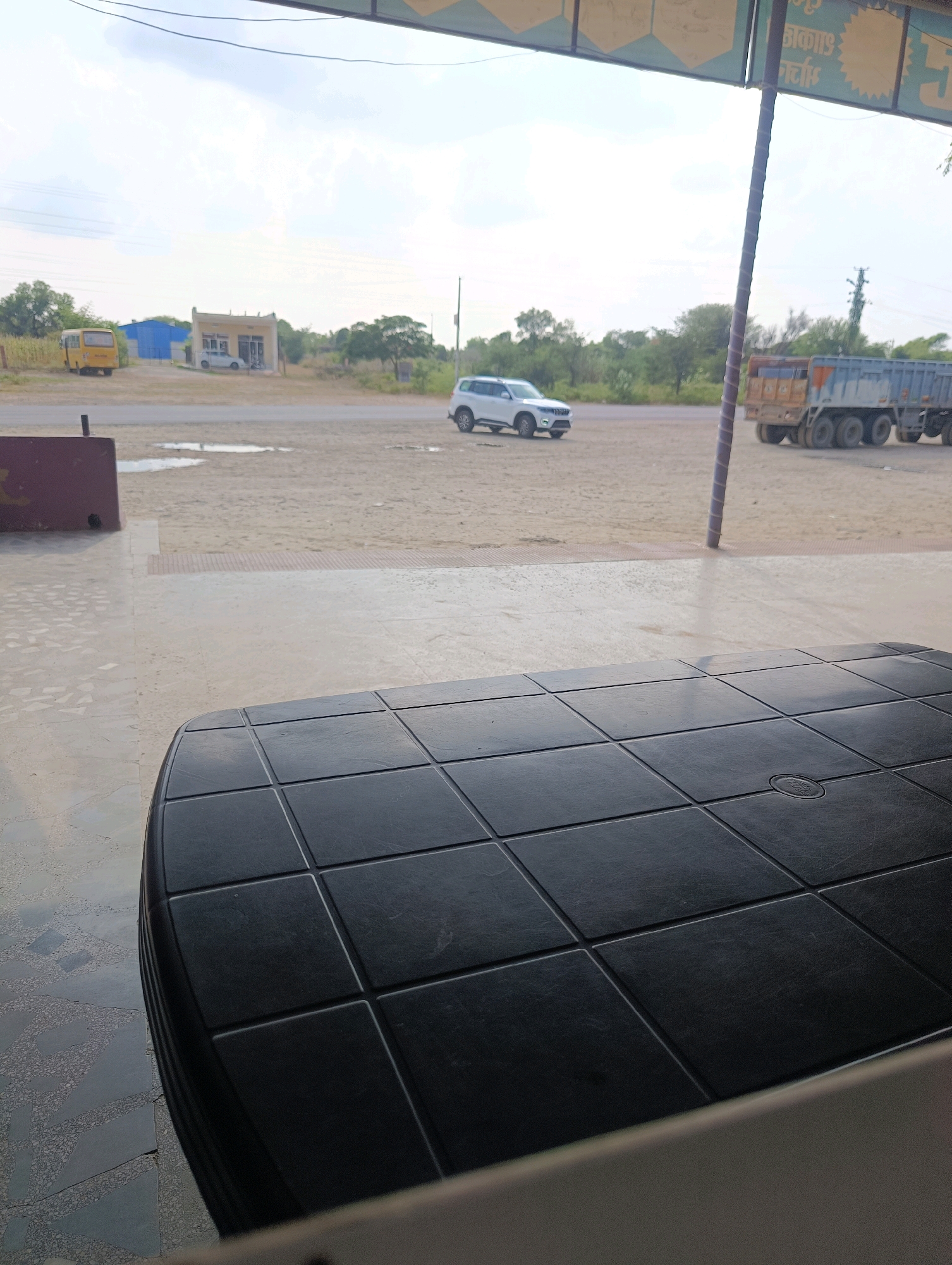 This image depicts a scene in Chhapri Bari, India. In the foreground, a black, square-patterned plastic table occupies the lower half of the frame. Beyond the table, the ground is paved with a light-colored, speckled material, transitioning to a dusty, gravelly area. A maroon barrier is visible to the left.
In the middle ground, a white SUV is driving on a paved road. Further in the distance, a yellow school bus is parked near a small building with a light-colored facade and a blue structure behind it. To the right of the road, a large, blue and orange truck with multiple axles is parked, its trailer loaded. Trees and vegetation are sparsely scattered throughout the landscape. The sky is overcast with light clouds. The overall impression is one of a roadside setting, possibly a rest stop or a commercial area, in a rural or semi-rural environment.