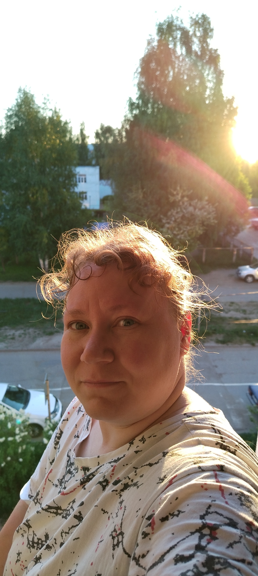 A middle-aged woman with light skin, short curly red hair, and green eyes is taking a selfie outdoors. She's wearing a patterned t-shirt. Behind her, there's a street with parked cars, trees, and a multi-story building. The sun is shining brightly, creating a lens flare effect in the upper right corner. The image was taken in the city of Chaikovsky, Russia.