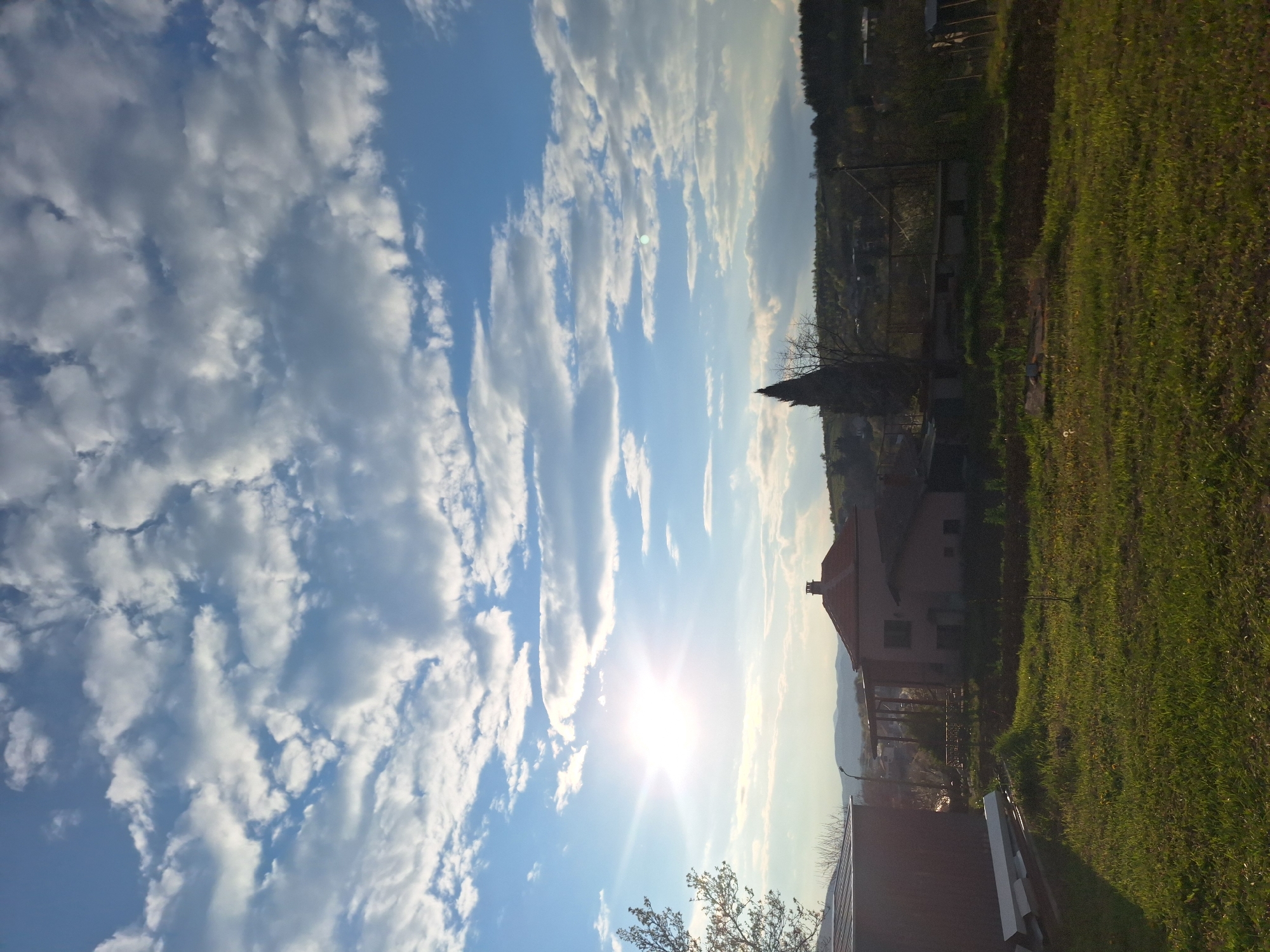 The image depicts a landscape scene with a clear blue sky partially covered by white, puffy clouds. The sun shines brightly, creating a glare. The right side of the image shows a green field with a house. In the background, a mountain ridge and trees can be seen. The scene appears to be in a suburban or rural setting. The location is Unknown, Unknown.