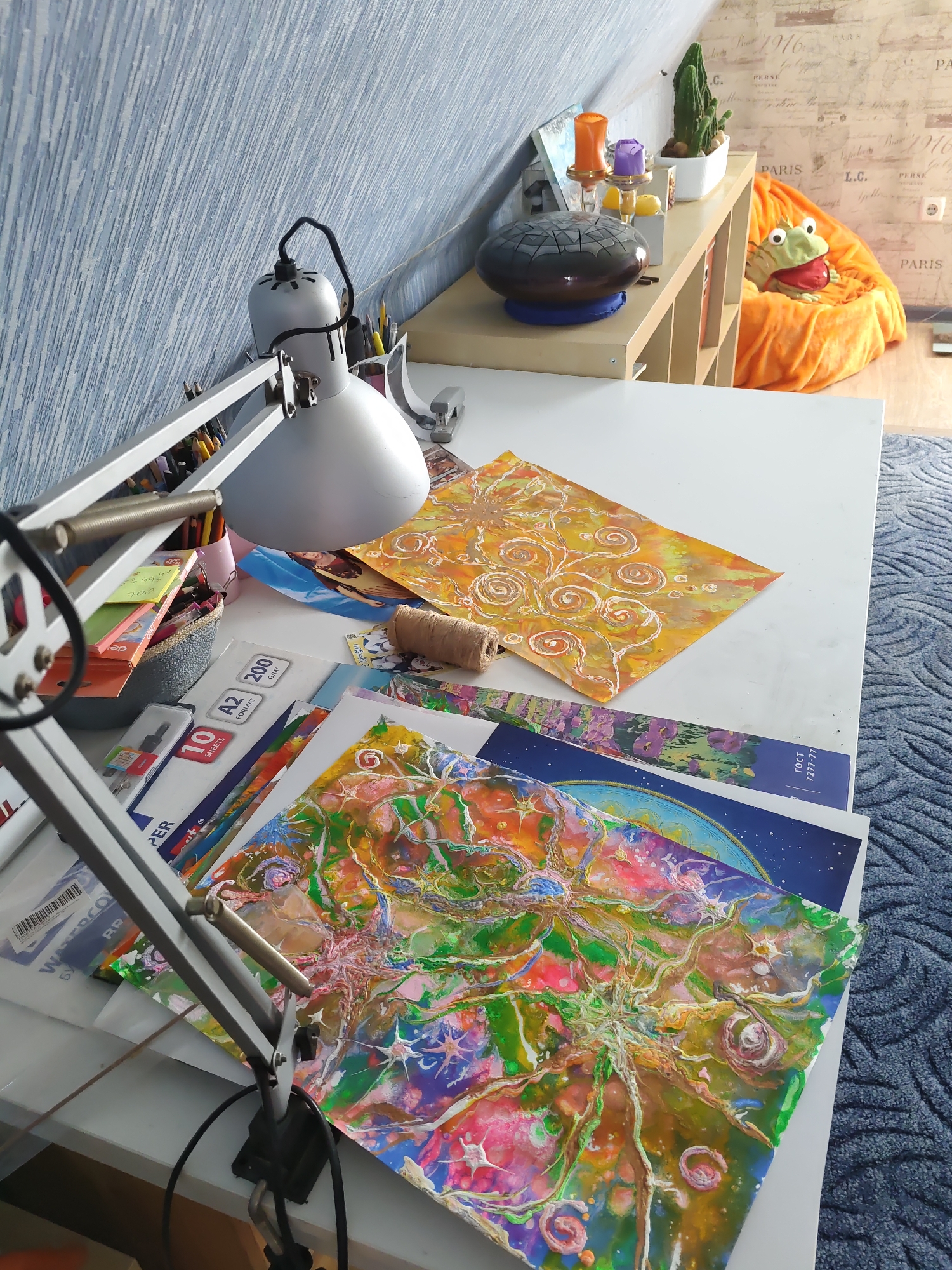 The image depicts an indoor scene, likely an artist's workspace, with a desk covered in art supplies and various papers. The desk is positioned against a slanted wall with blue textured wallpaper. On the desk, there is a silver desk lamp, a basket filled with art supplies like pens and pencils, stacks of paper including A2 format paper, and several pieces of artwork. One piece is a yellow paper with an orange swirl pattern, and another is a vibrant, abstract painting with green, pink, and yellow colors. A roll of twine is also visible on the desk.
To the right of the desk, there is a shelf unit with several items on it, including a glass object with colored lights, a potted cactus, and a plush frog toy sitting in an orange beanbag chair. The background wall has a map-like wallpaper with text that appears to include "PARIS".
Although the user prompt specifies a location in Rostov-on-Don, Russia, there are no direct visual cues within the image to confirm this specific location. The scene is characteristic of a creative studio or a well-organized home art area.