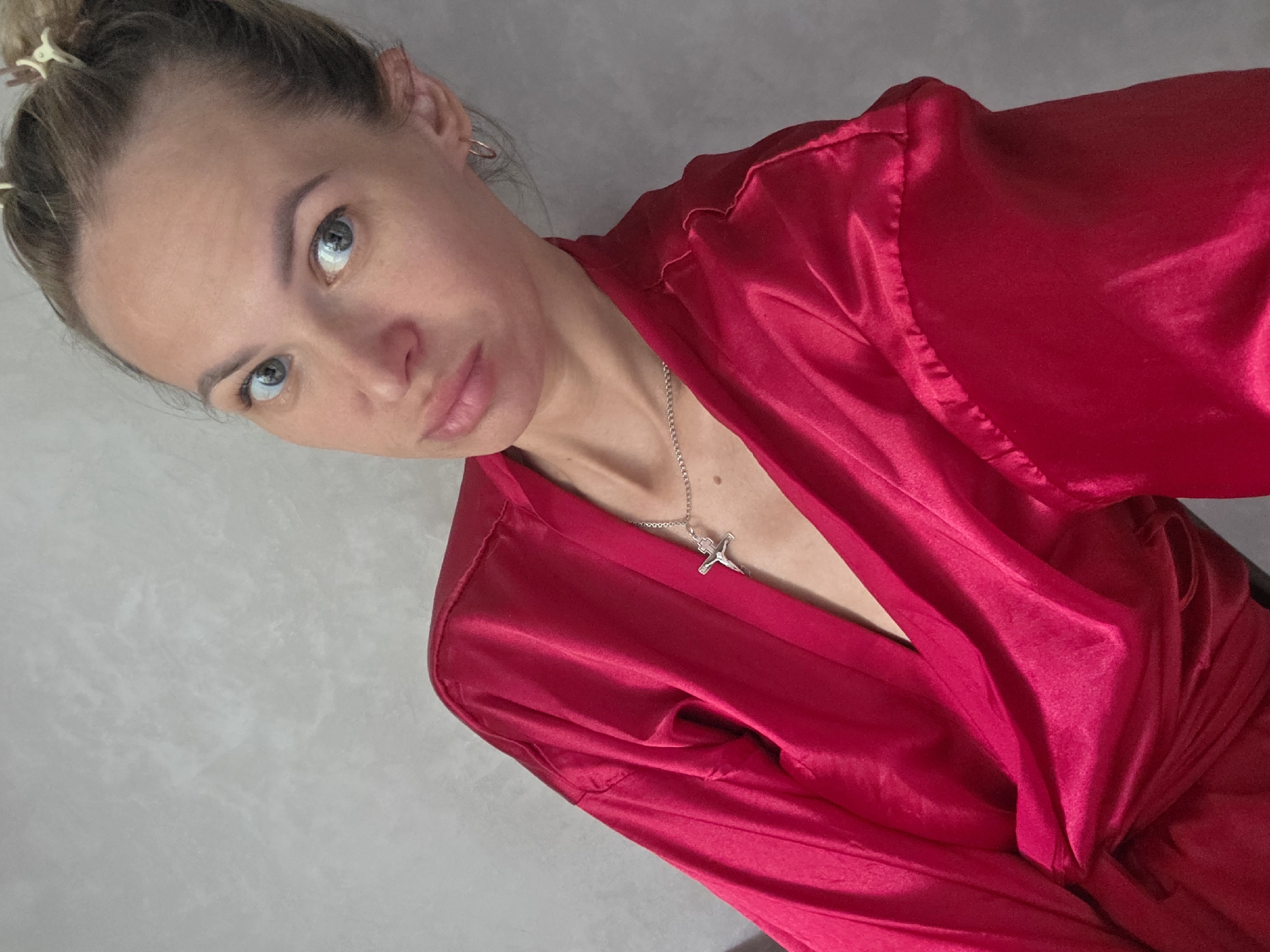 A woman with light blonde hair pulled back is looking at the camera. She has light blue eyes and a somewhat pouty expression. She is wearing a bright red satin robe and a silver cross necklace. The background appears to be a light grey wall. This photo was taken in Chaikovsky, Russia.