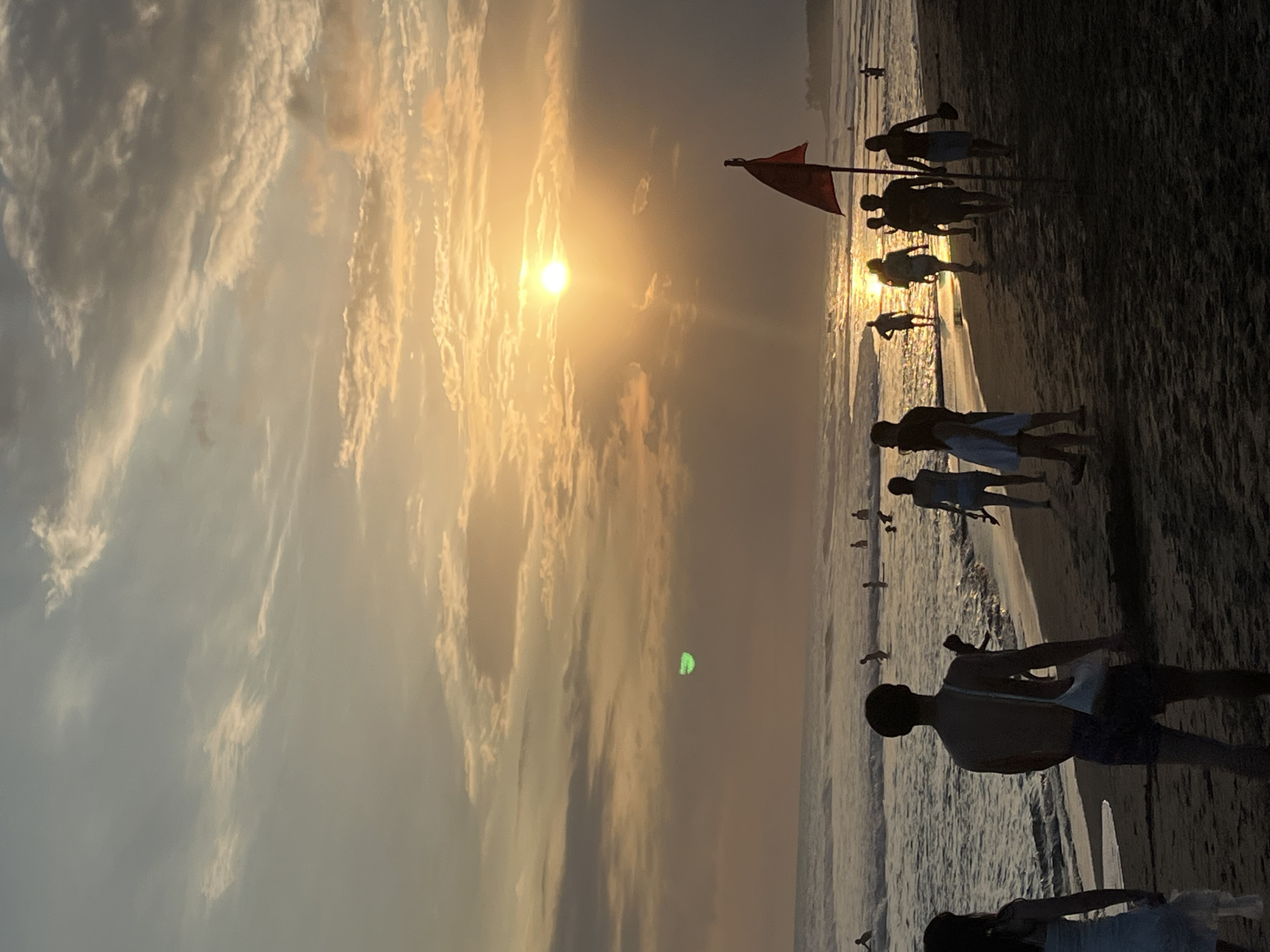 This is a medium shot photograph of a beach scene at sunset in North Kuta, Indonesia. The sun is low on the horizon, casting a warm glow across the sky and reflecting off the ocean waves. Several people are present on the beach, some walking along the shore, others wading in the water. A red flag on a pole stands prominently on the sand, suggesting a designated swimming area or a warning. The sand appears dark and wet near the water's edge, with dry sand further up the beach. The sky is filled with a mixture of clouds, some illuminated by the setting sun. There is a faint green circular anomaly visible in the upper portion of the sky, which could be a lens flare or other photographic artifact.