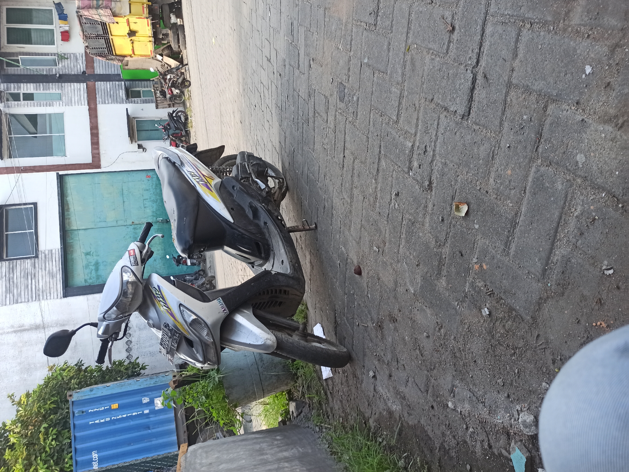 A silver Yamaha Mio scooter is parked on a paved sidewalk in front of a building with light-colored walls and a large green metal door. The scooter is angled towards the viewer, with its front wheel turned slightly to the left. A license plate with Indonesian characters is visible on the front of the scooter. To the left of the scooter, a blue shipping container with white text is partially visible, and a large green planter with lush green foliage stands beside it. In the background, a yellow truck with a covered cargo area is parked, and several other motorbikes are visible further down the street. The ground is a mixture of dirt and paving stones, with some debris scattered around. The lighting suggests it is daytime.