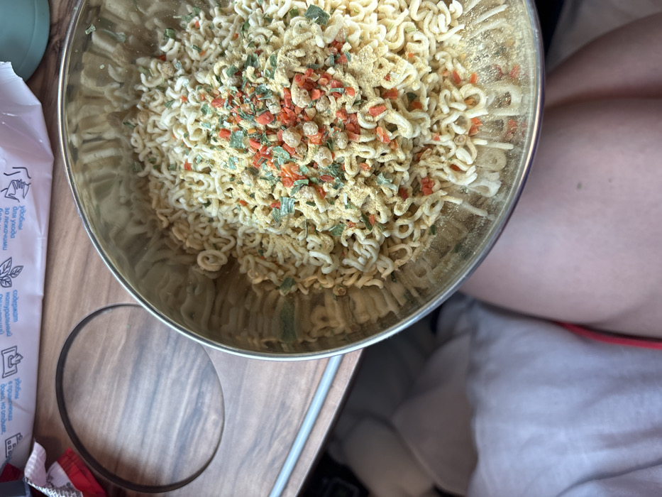The image depicts an overhead view of a silver metallic bowl filled with uncooked instant noodles and seasoning, resting on a wooden-patterned fold-down tray table. The scene is located within a passenger train, likely in or passing through Tatarsk, Russia, as indicated by Cyrillic text visible on a white package or leaflet in the background.
The instant noodles are pale yellow and have a characteristic wavy, dehydrated form. They are generously covered with a dry seasoning mix comprising yellow powder, small green flakes (likely dried herbs), and orange-red flakes (possibly dried carrots or bell peppers), along with some light beige granular pieces.
The tray table features a prominent circular indentation, typically for holding a cup or bottle. A thin, grey metallic bar is visible running horizontally along the left edge of the tray. In the upper background, a white rectangular object displays various icons and Cyrillic text, including phrases such as "уход за руками" (hand care) and "содержит витамины" (contains vitamins), suggesting a personal hygiene product or informational material.
The lower portion of the image reveals the upper thighs of a person, indicating a first-person perspective. One leg is partially covered by light-colored fabric, while the other is bare, showing skin. A small strip of red fabric is visible between the legs, suggesting part of the person's clothing. No direct actions or interactions are depicted, only the static arrangement of objects. The environment is consistent with a train car interior during a journey.