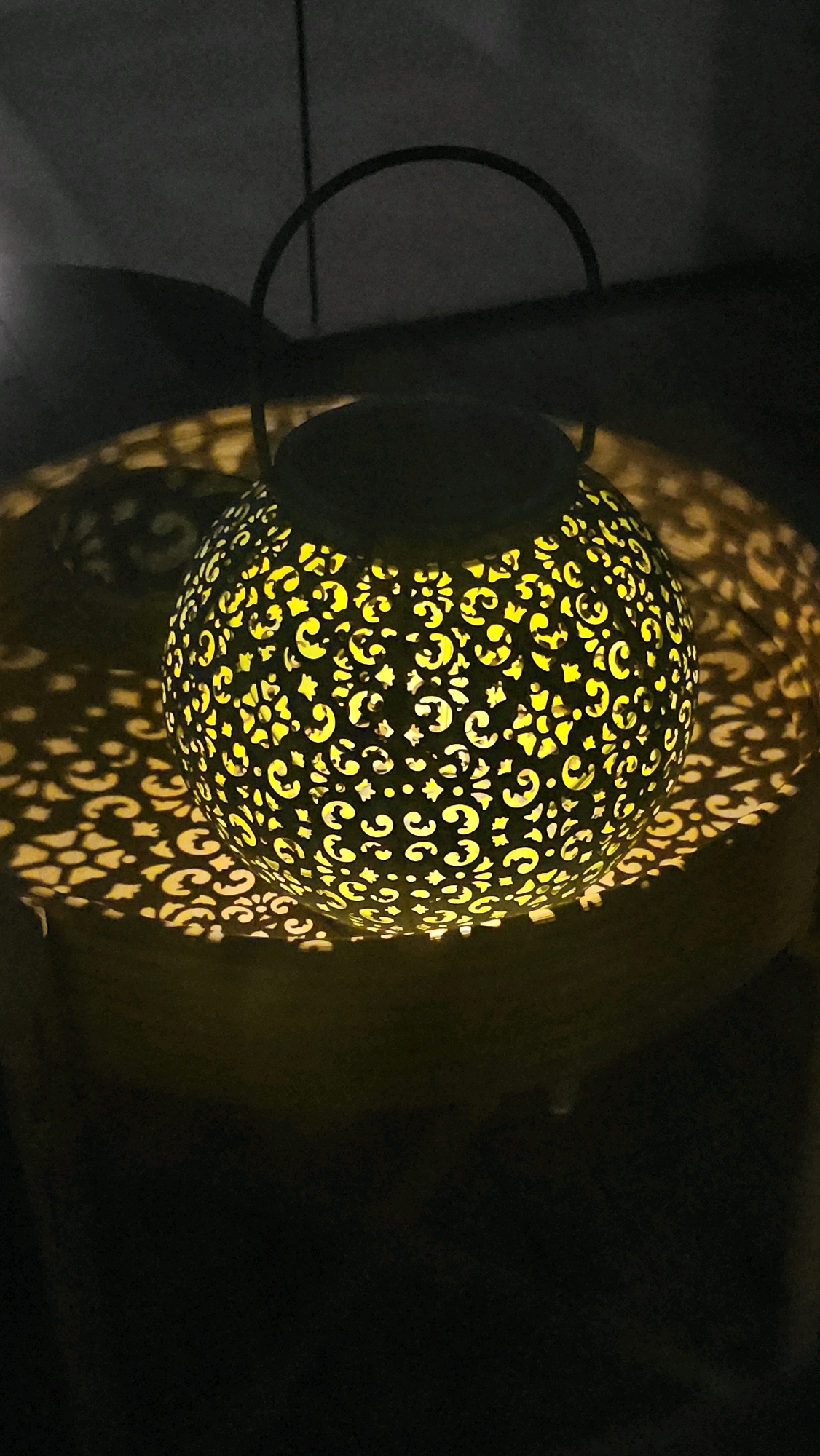 The image shows a decorative lantern with intricate cutouts, casting a patterned shadow on a dark surface. The lantern is spherical and emits a warm yellow light from within, highlighting its ornate design. The background is dark and out of focus, suggesting it is nighttime or indoors with low lighting. There is no indication of Cologne, Germany in the image.