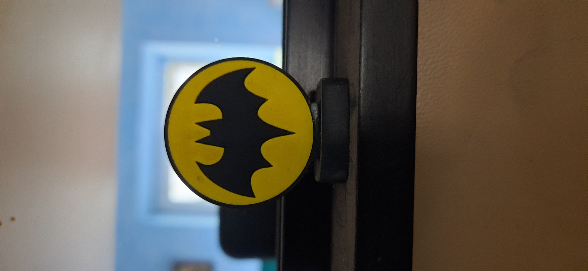 A close-up, eye-level shot shows a black and yellow Batman logo on a small black base. The logo is circular with the iconic bat silhouette in black against a yellow background. The base is a small, rectangular block. The objects are positioned on a dark, horizontal surface that appears to be a shelf or countertop. In the blurred background, a wall painted light blue is visible, with what seems to be a window or doorway with white trim. There are no people visible in the image. The scene is indoors, in a room. The location is not specified as Dnipro, Ukraine.